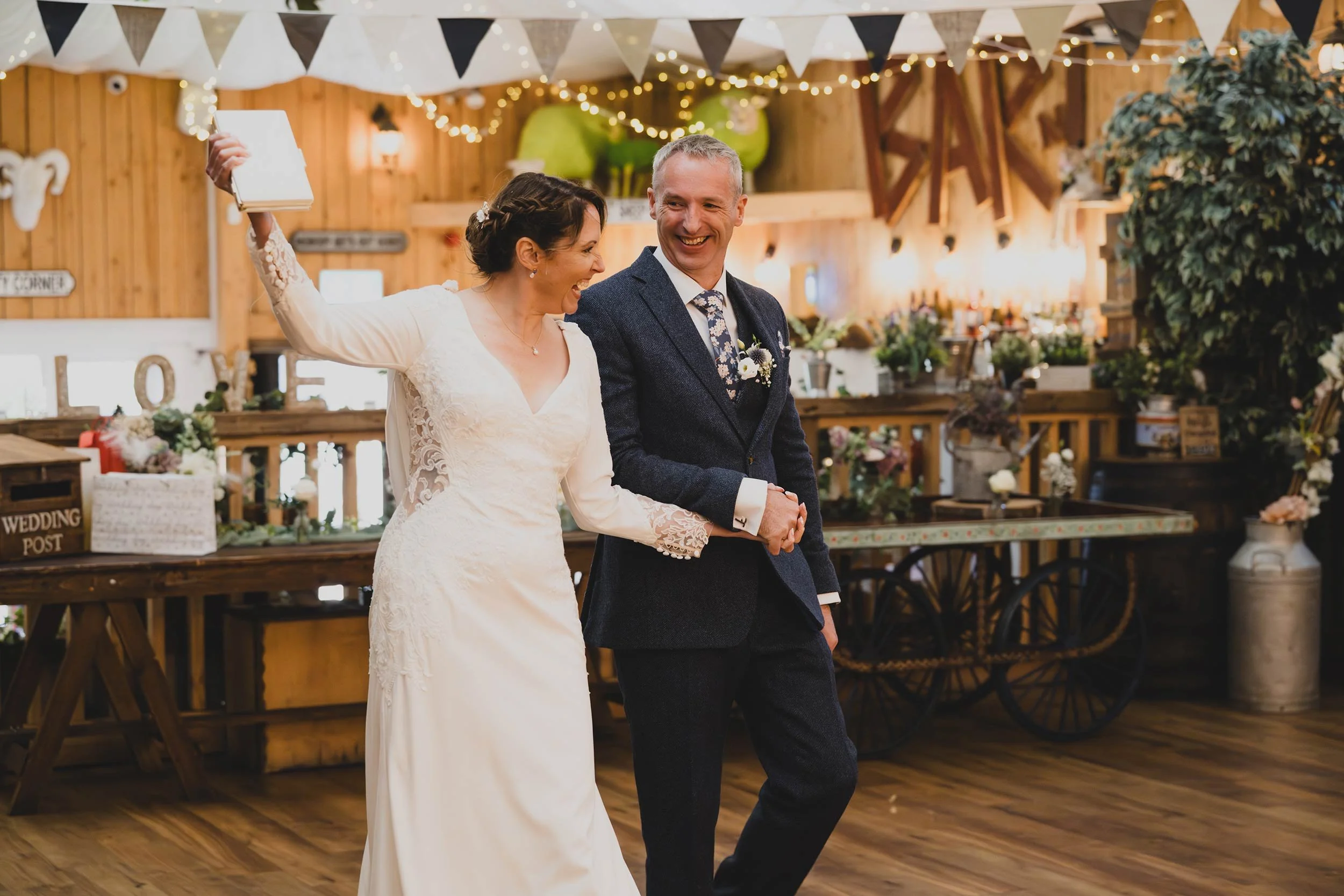 A bride and groom dancing and smiling at their wedding reception, decorated with string lights, bunting, balloons, and floral arrangements in a rustic barn setting.