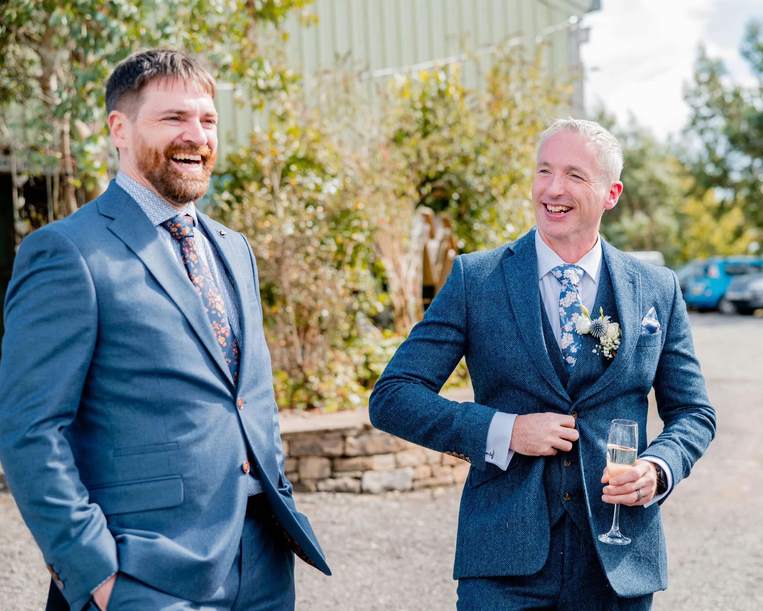 Two men in suits smiling and talking outdoors during a celebration, with one holding a glass of champagne.