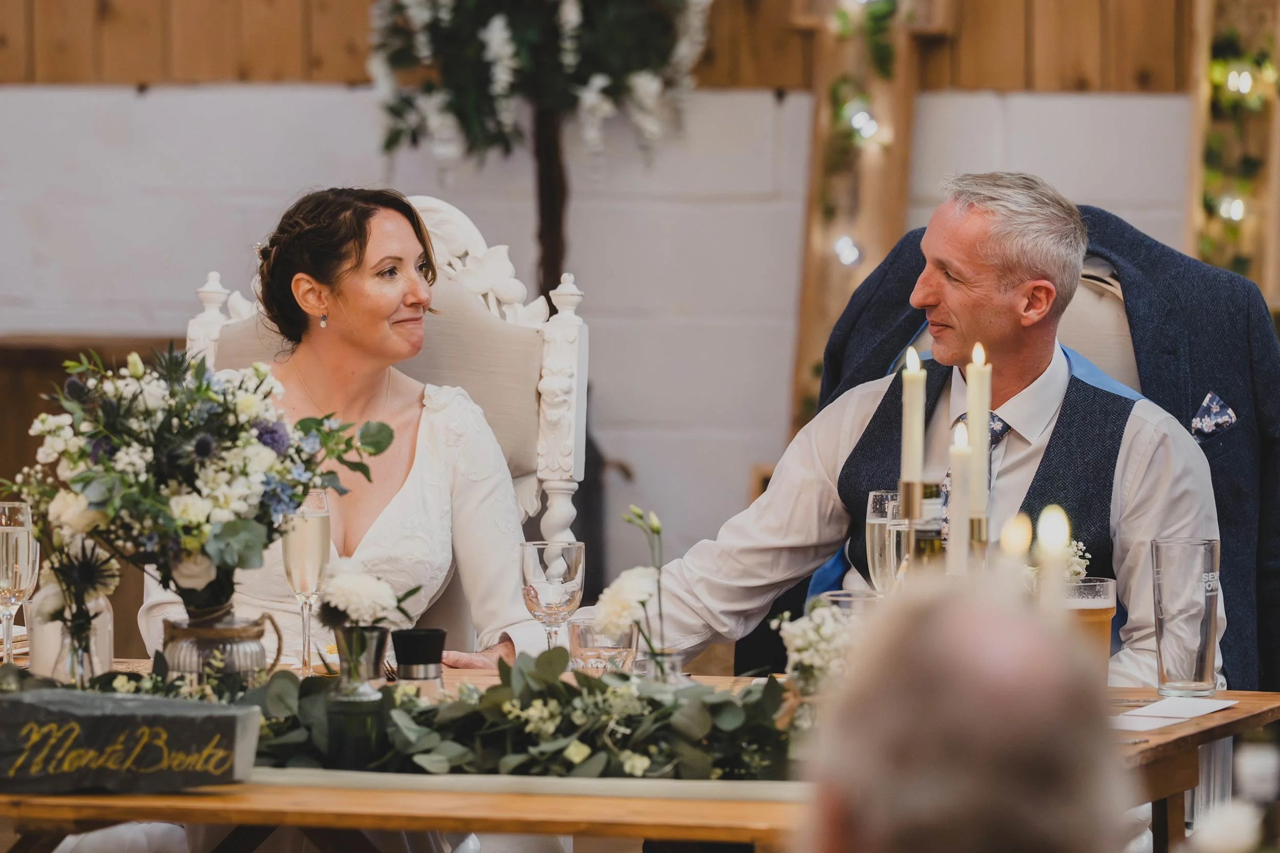 A wedding reception with a bride and groom sitting at a decorated table, holding hands and gazing at each other. The table has floral arrangements and lit candles. The bride is wearing a white dress, and the groom is in a white shirt with a dark vest
