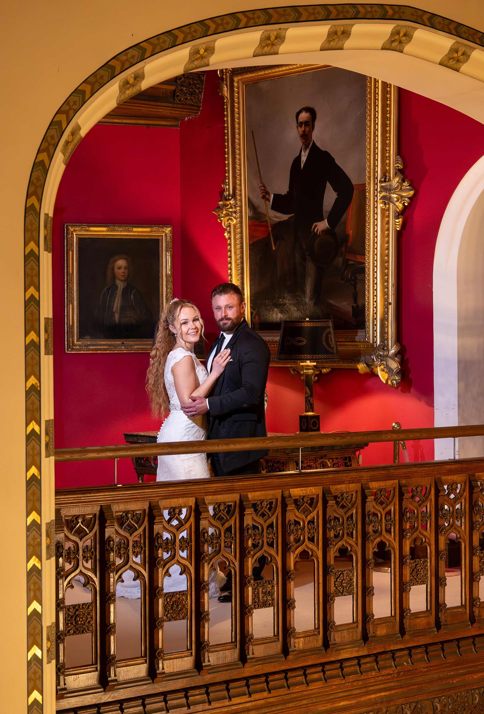 A bride and groom dancing on a balcony inside a museum or historical building, with framed portraits on a red wall behind them, ornate wooden railing, and gold framed large portrait of a man holding a stick.
