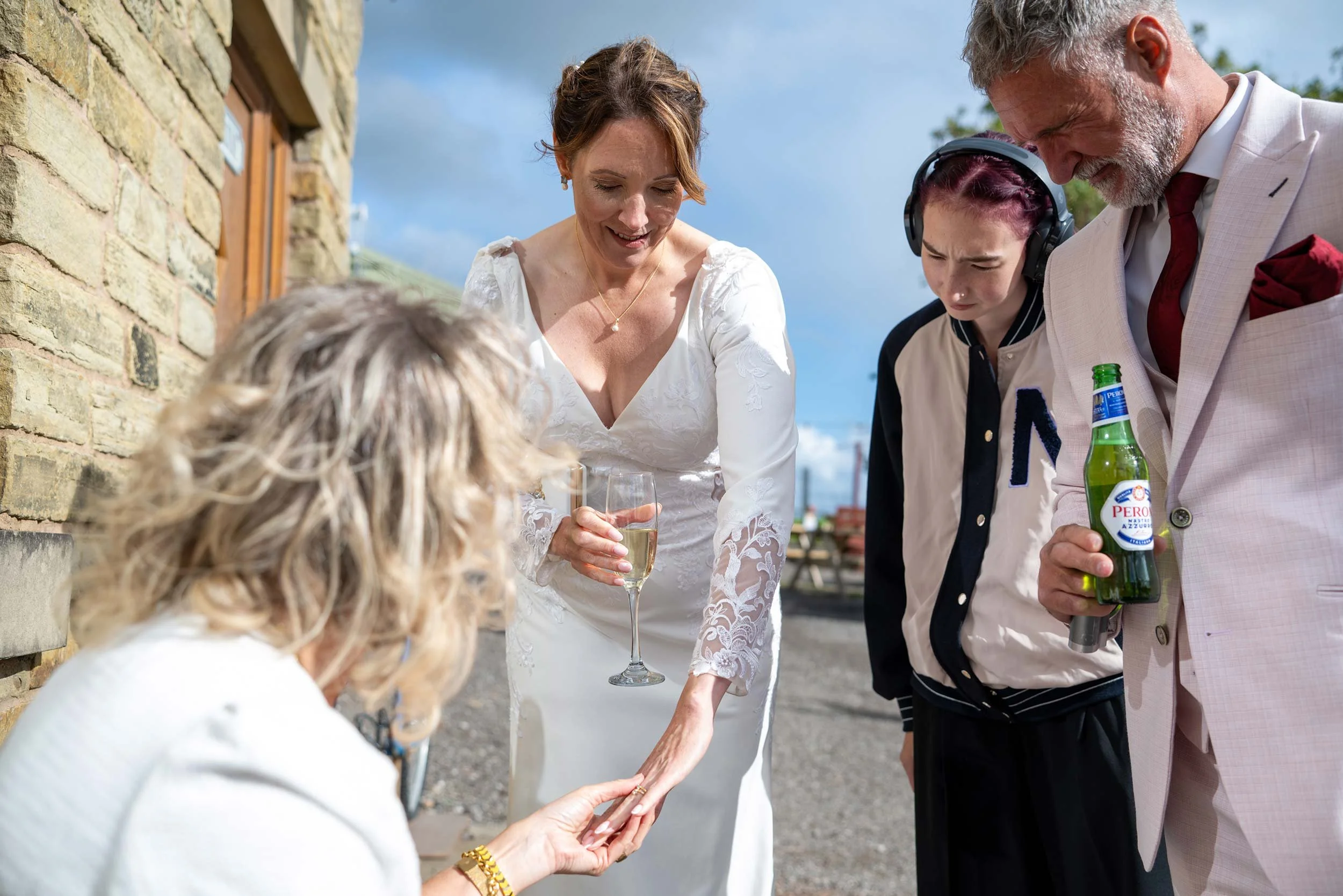A woman in a white dress is being presented with a ring during an outdoor ceremony, surrounded by four people, including a woman with a glass of champagne and a man holding a beer, under a partly cloudy sky.