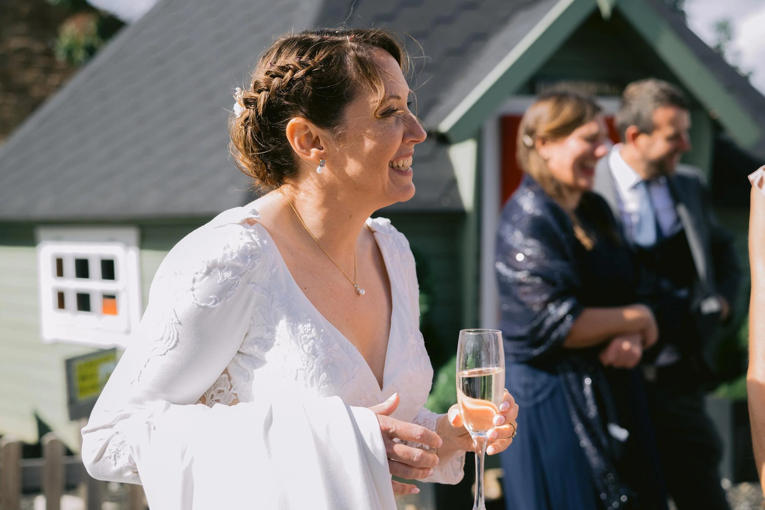 A woman in a white dress holding a glass of champagne, smiling at an outdoor celebration with people and a house in the background.