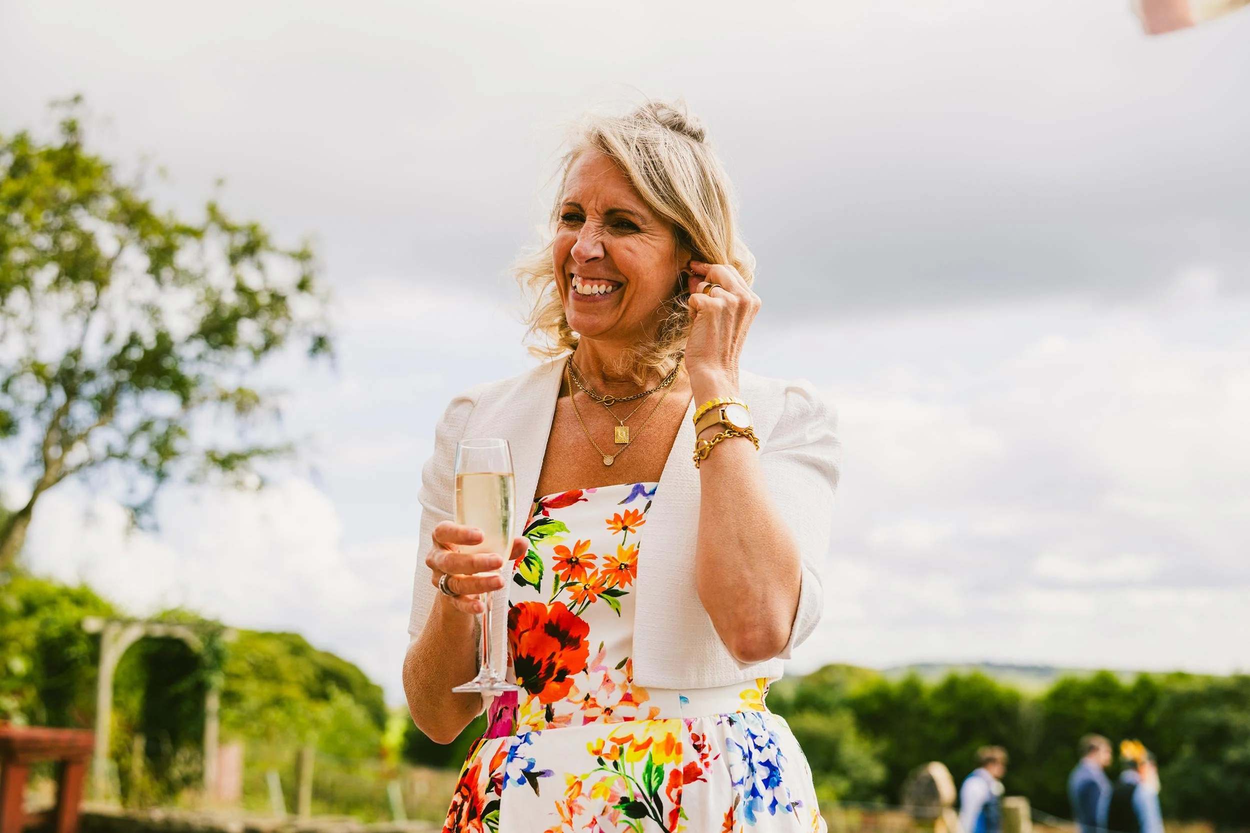 A smiling woman with curly blonde hair, wearing a white blazer and a floral dress, holds a glass of champagne at an outdoor event.