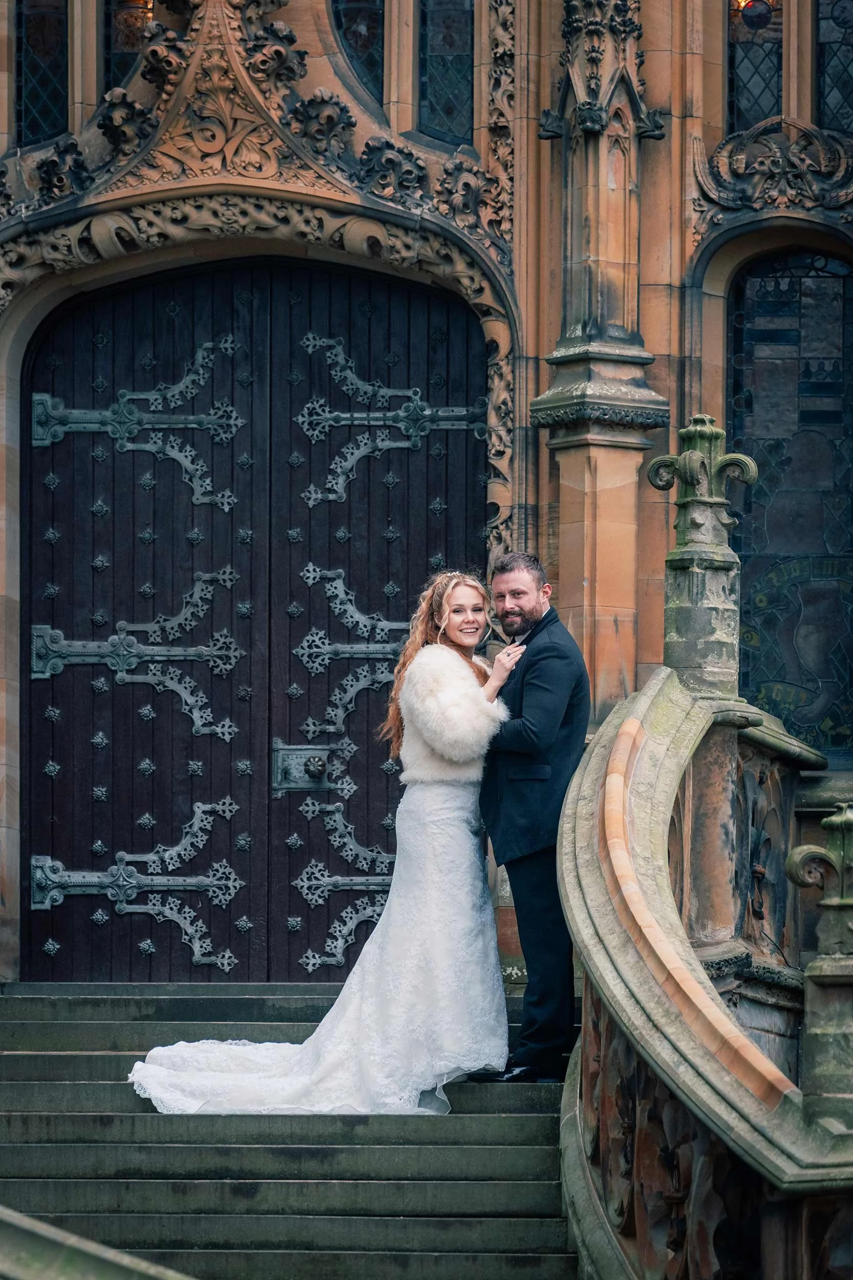 A bride and groom standing on stone steps in front of a gothic-style building with intricate carved details and large doors. The bride has long, wavy red hair, and is wearing a white lace wedding dress and a fur shawl. The groom has dark hair and a b