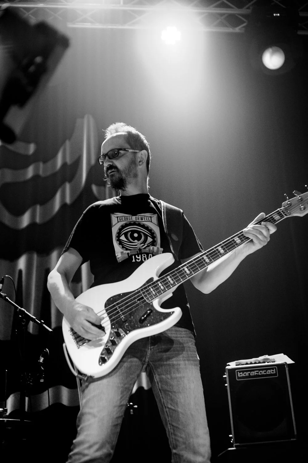 A man playing an electric bass guitar on stage, wearing glasses and a graphic t-shirt, with stage lights overhead and curtains in the background.