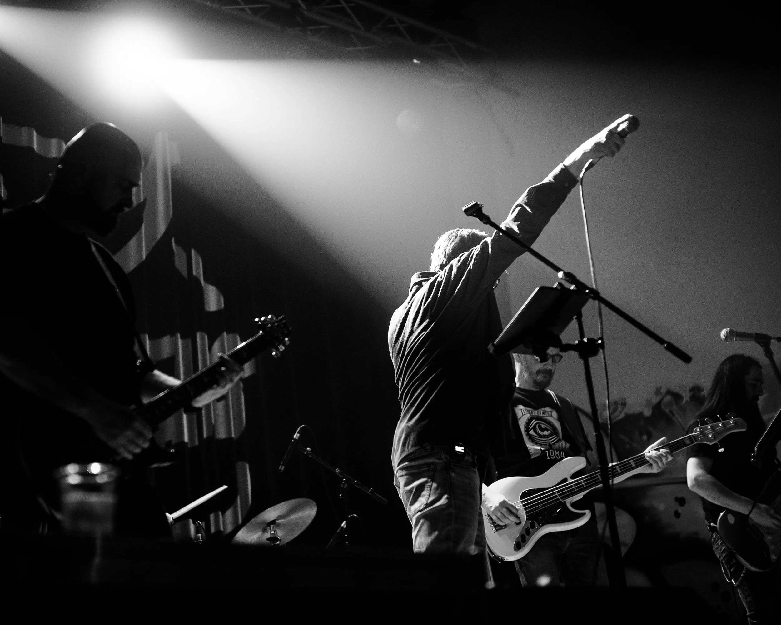Black and white photo of a band performing on stage, with one member raising a hand and holding a microphone, others playing guitars, in a concert setting.