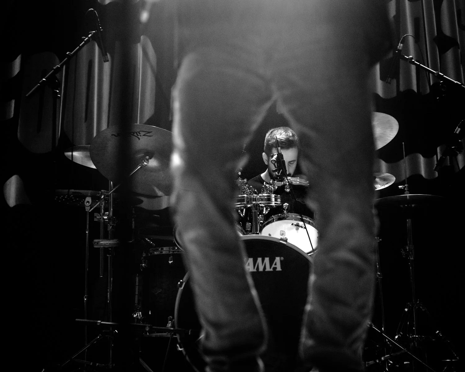 A black and white photo of a drummer performing on stage, seen through the legs of a person standing in front of him.