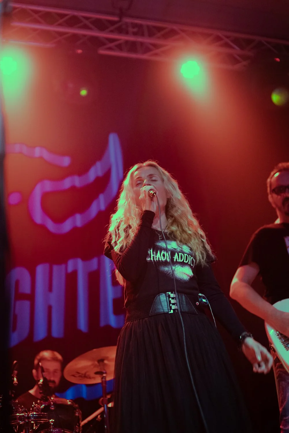 A woman with long curly blonde hair singing into a microphone on stage, with a band member playing drums in the background. The stage is lit with red and green lights, and a large screen behind displays a stylized image and text.