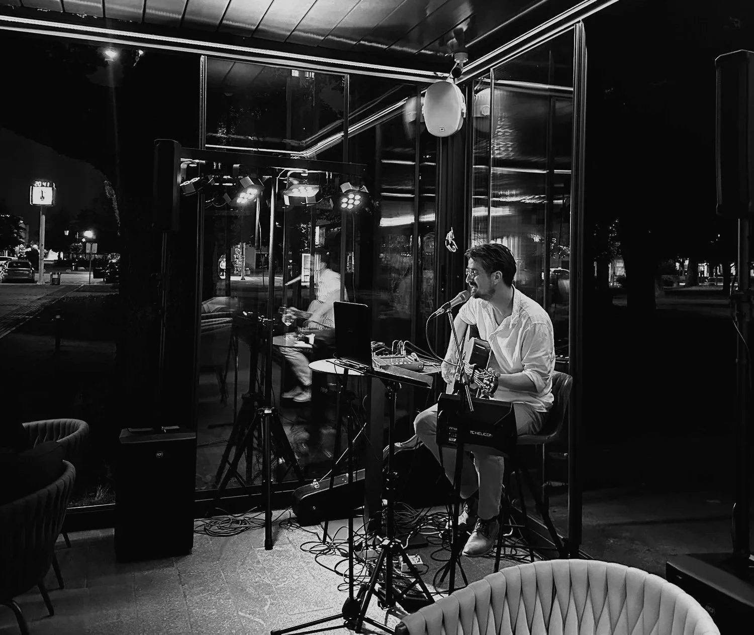 A musician in a white shirt sitting on a stool, playing guitar and singing into a microphone during a live performance at night, inside a glass-walled venue with reflections of trees outside.