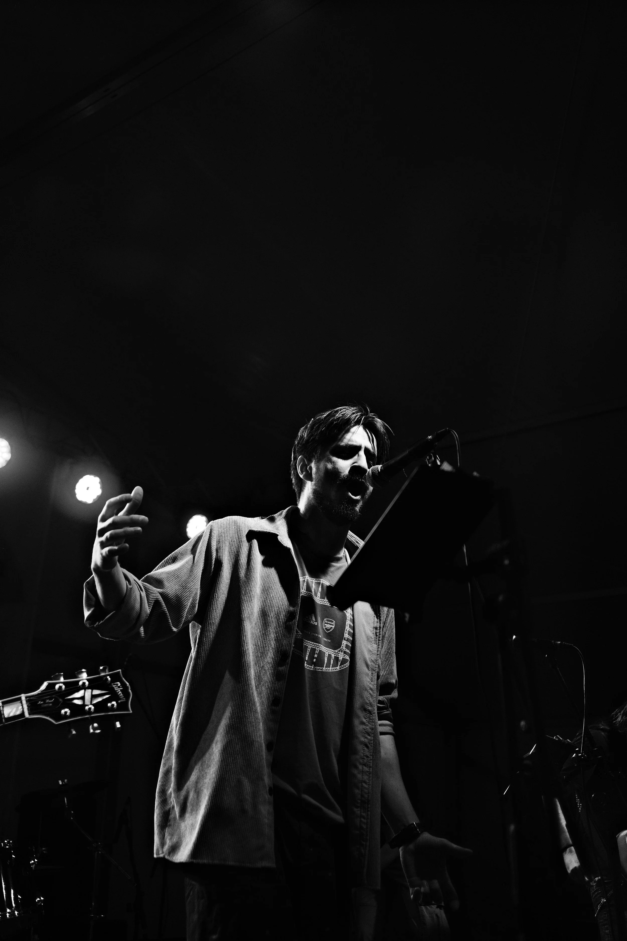 A black and white photo of a man singing into a microphone on stage, with music sheets in front of him and a guitar partially visible on the left.