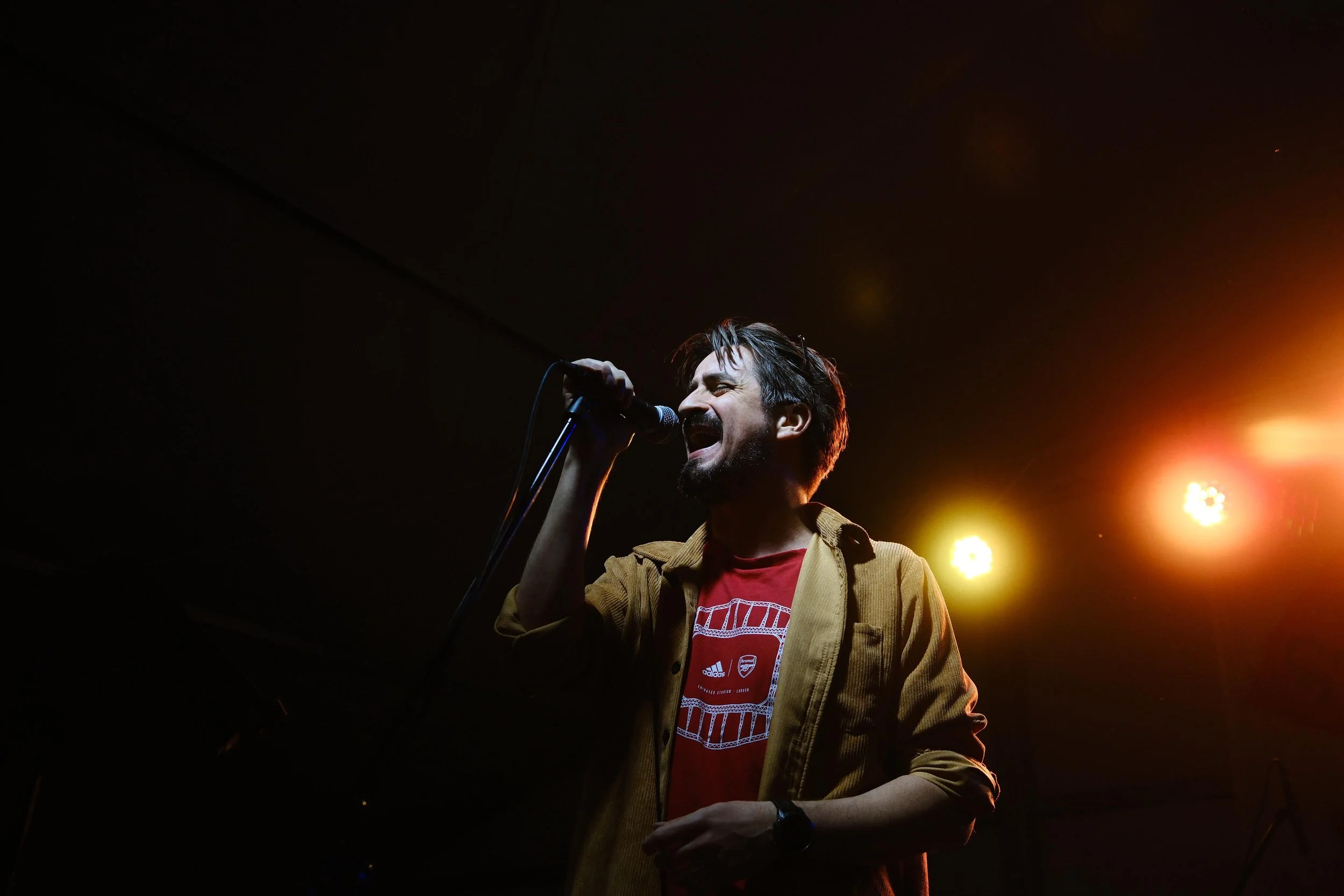 A man singing into a microphone on stage, illuminated by yellow and orange stage lights, wearing a red Adidas Arsenal shirt and a tan jacket.