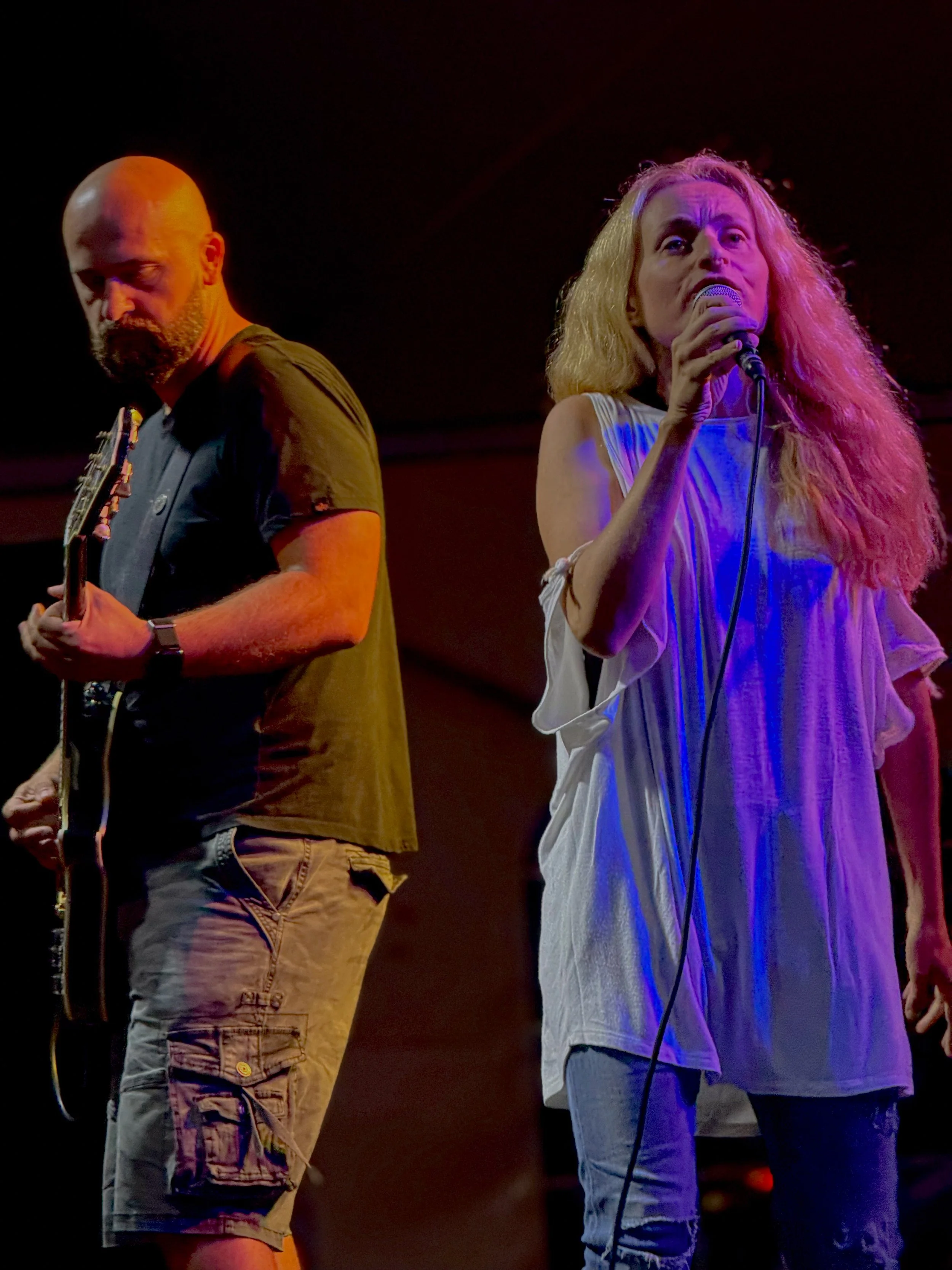 A man playing guitar and a woman singing into a microphone on stage with colored lighting.