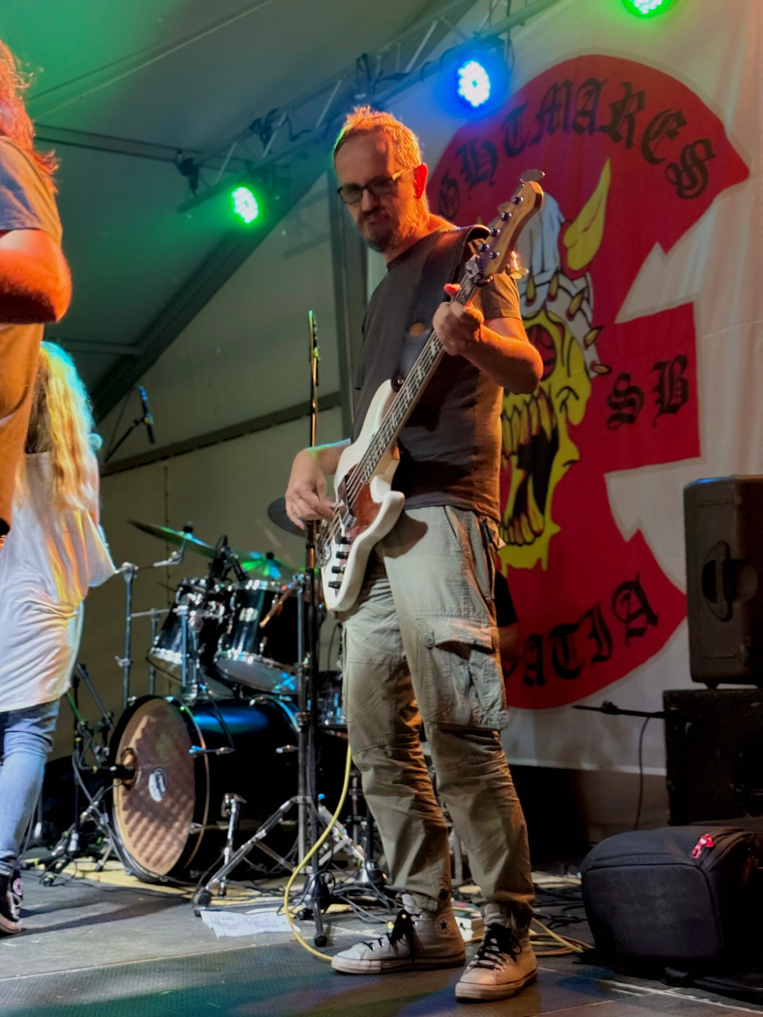 A man playing an electric bass on stage during a live music performance, with colorful stage lights and a drum set in the background, and a large red banner with a yellow knight's helmet and text behind him.