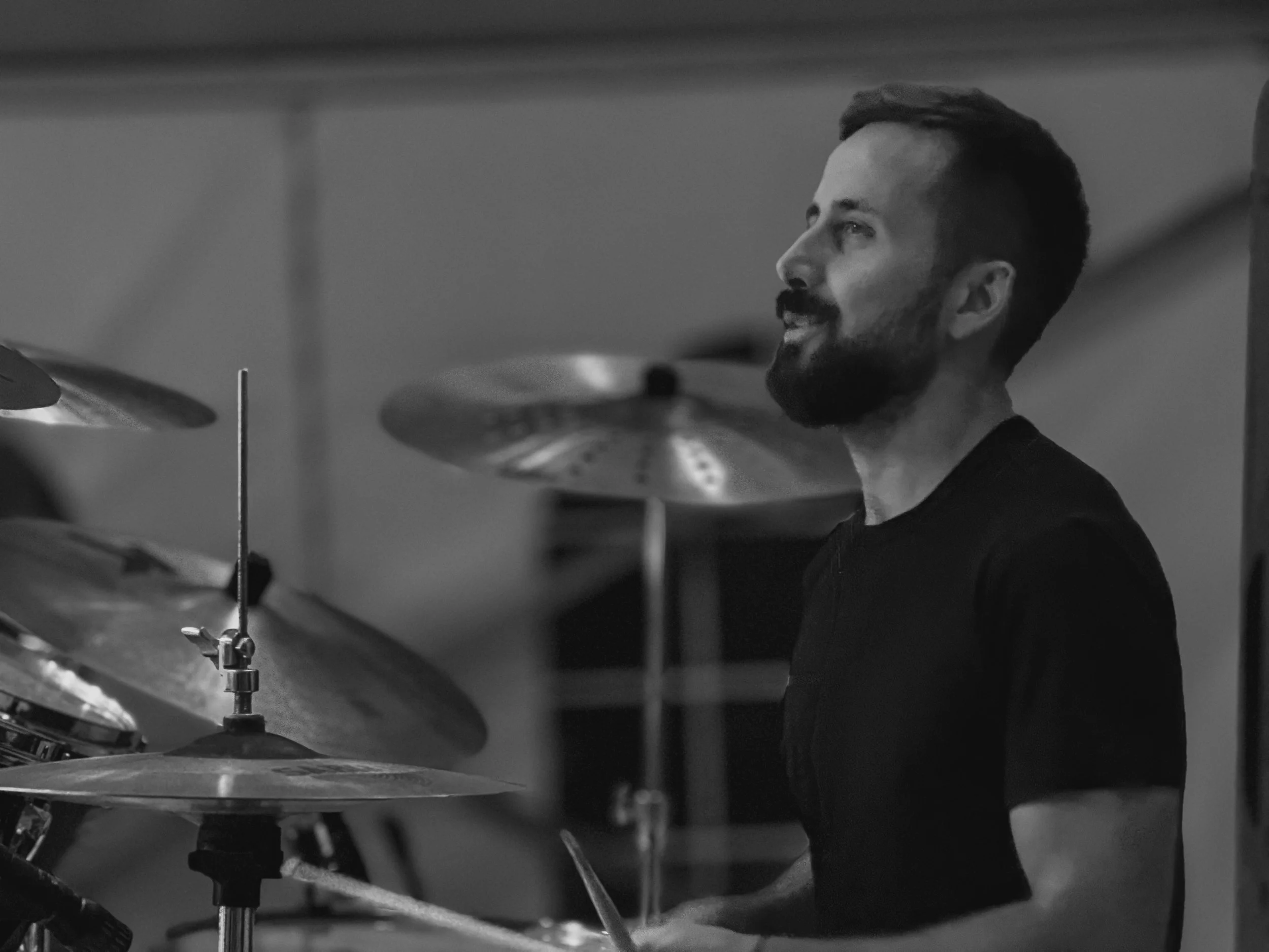 A man with a beard playing drums in a concert under tent, surrounded by cymbals and drums, smiling and facing to the right.