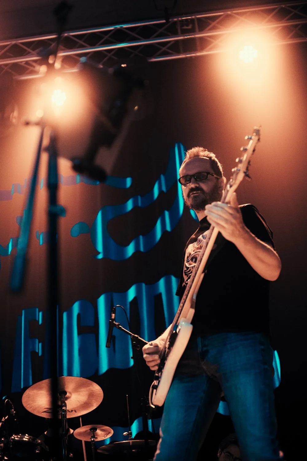 A man with glasses and a beard playing an electric guitar on stage, with warm stage lighting and a dark background with blue text.