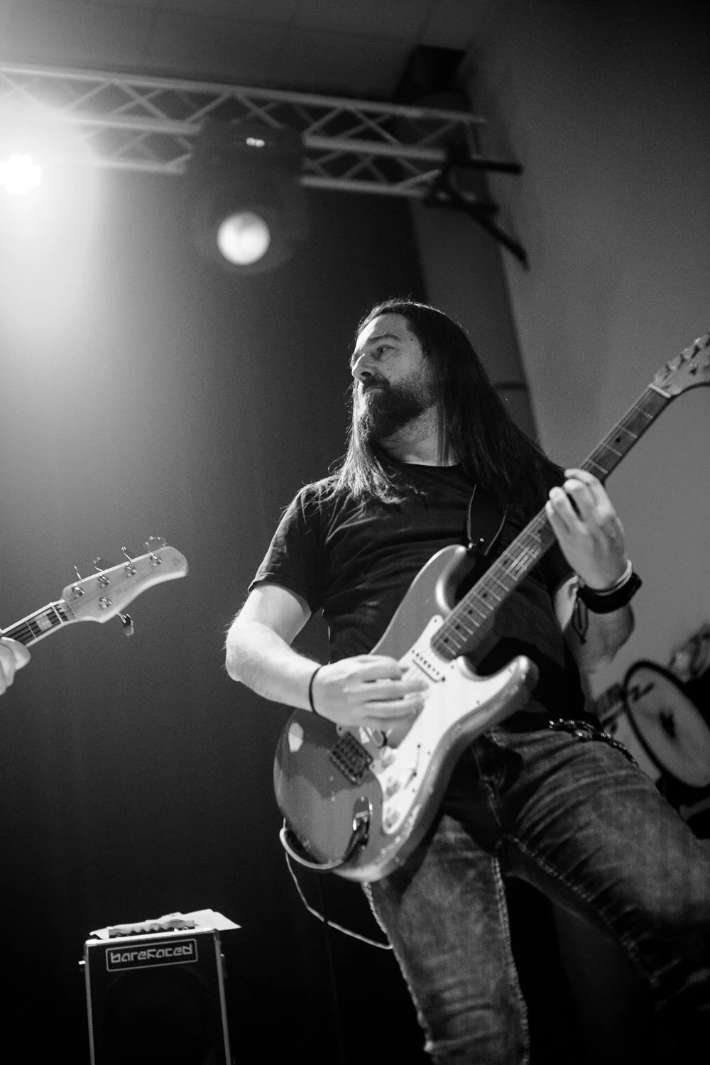 A man with long dark hair and a beard playing an electric guitar on stage in black and white.