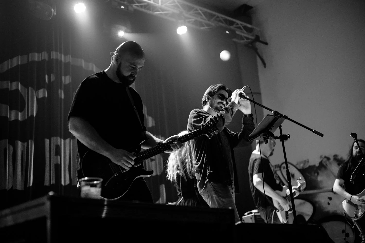 Black and white photo of a band performing on stage, with two guitarists and a singer, in a dimly lit venue with spotlights and curtains in the background.