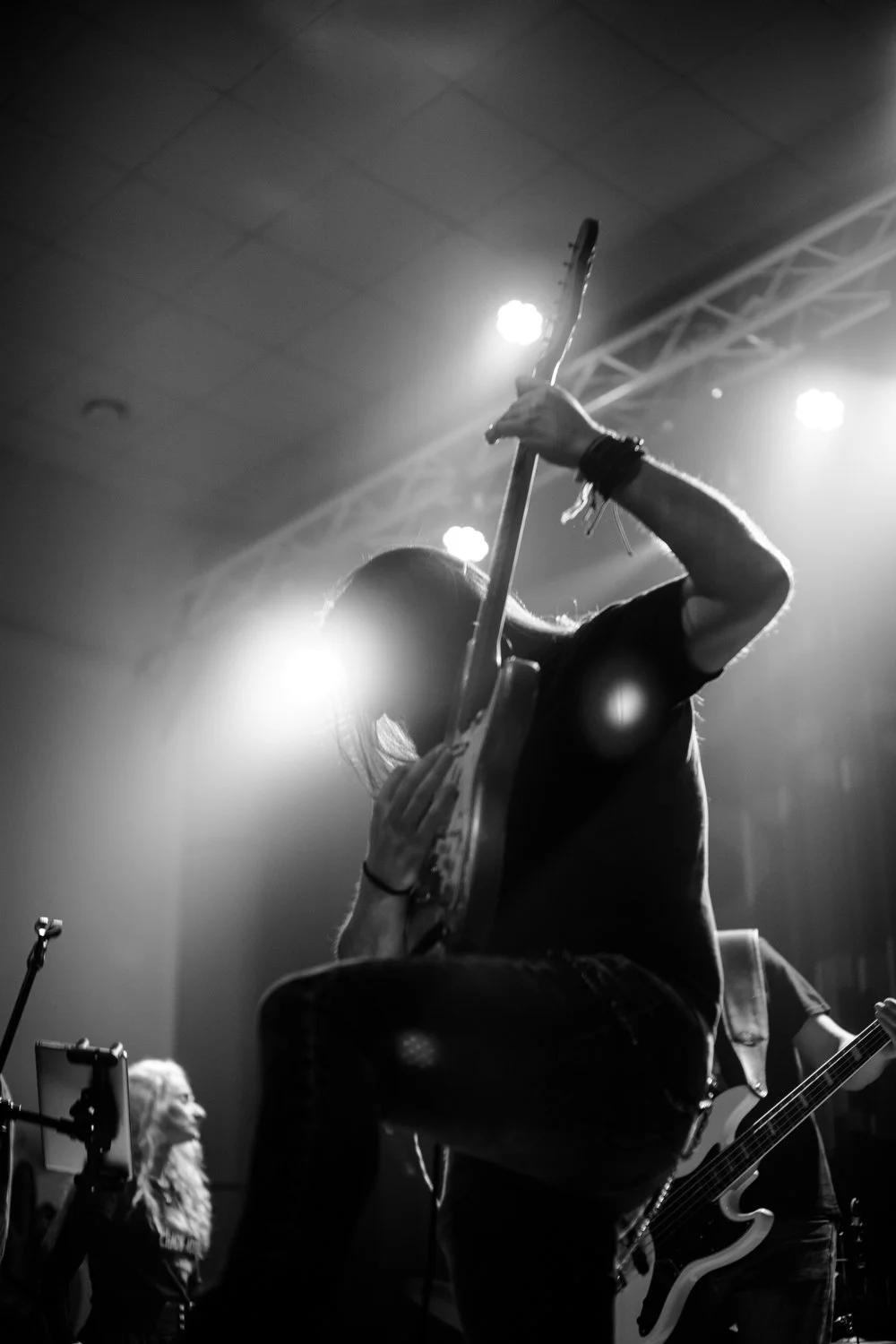 Silhouette of a guitarist on stage with bright lights and a singer in the background.