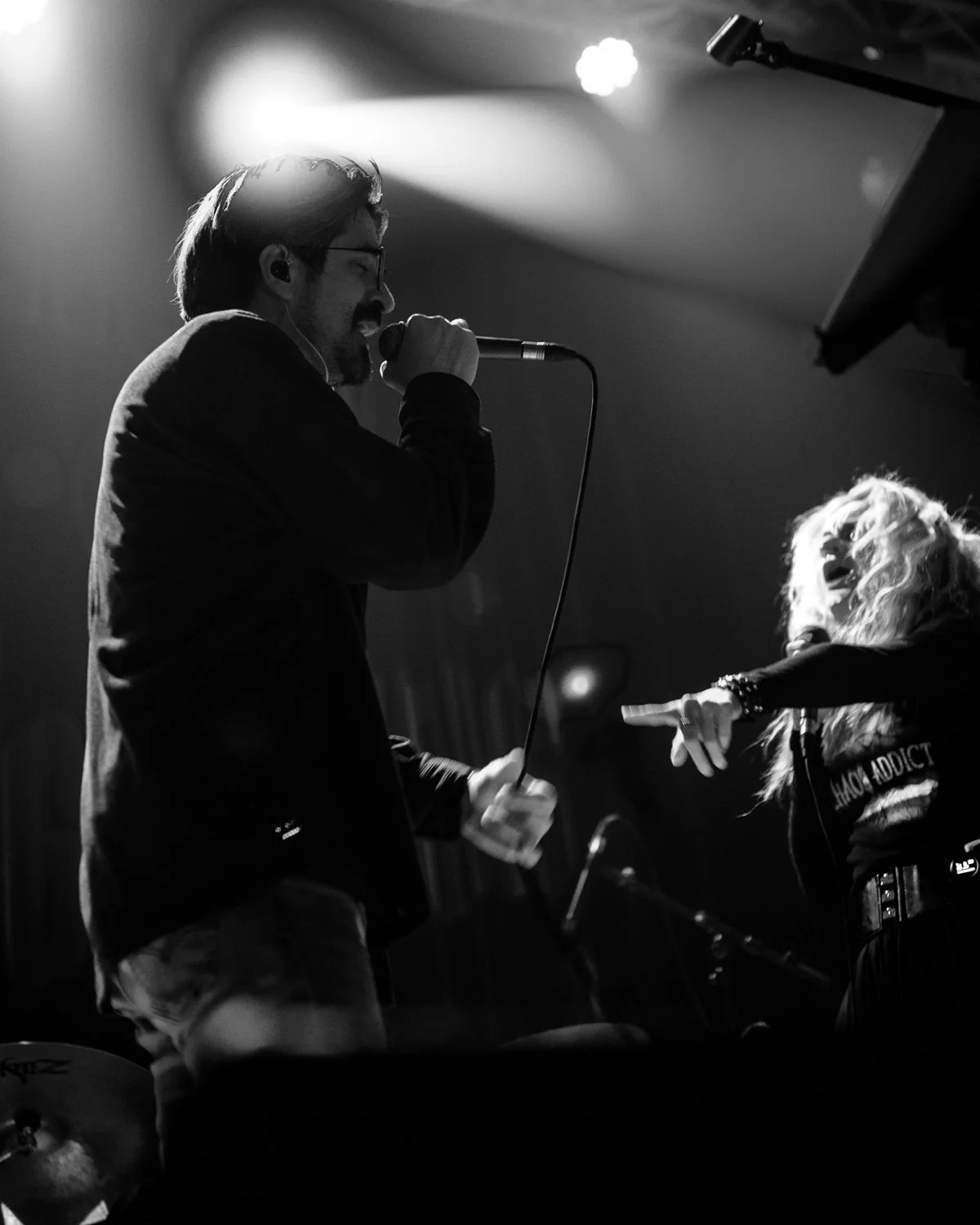 A man and a woman singing into microphones on stage, with stage lights overhead, in black and white.