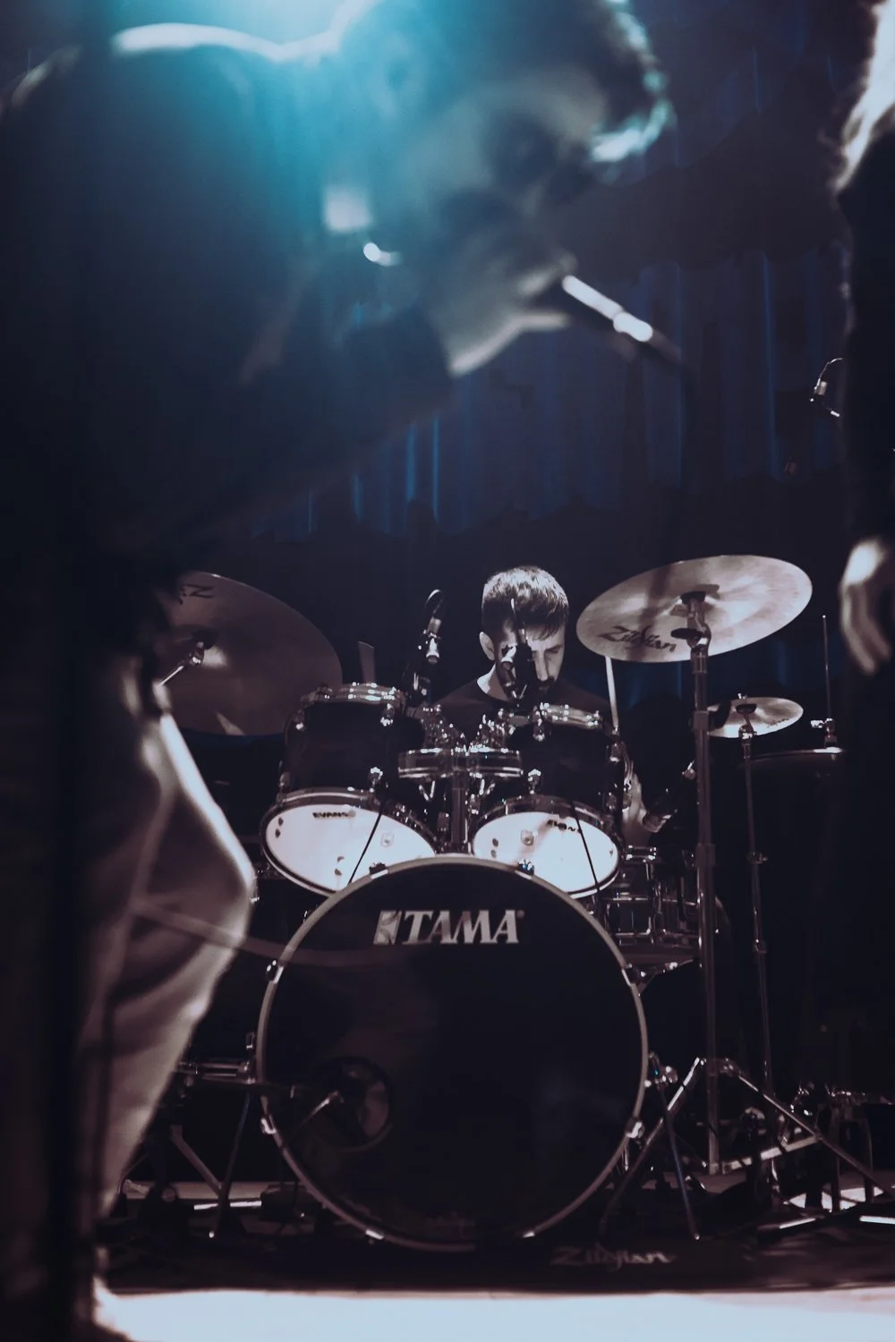 A drummer playing a TAMA drum set on stage, illuminated by stage lights, with a dark background.