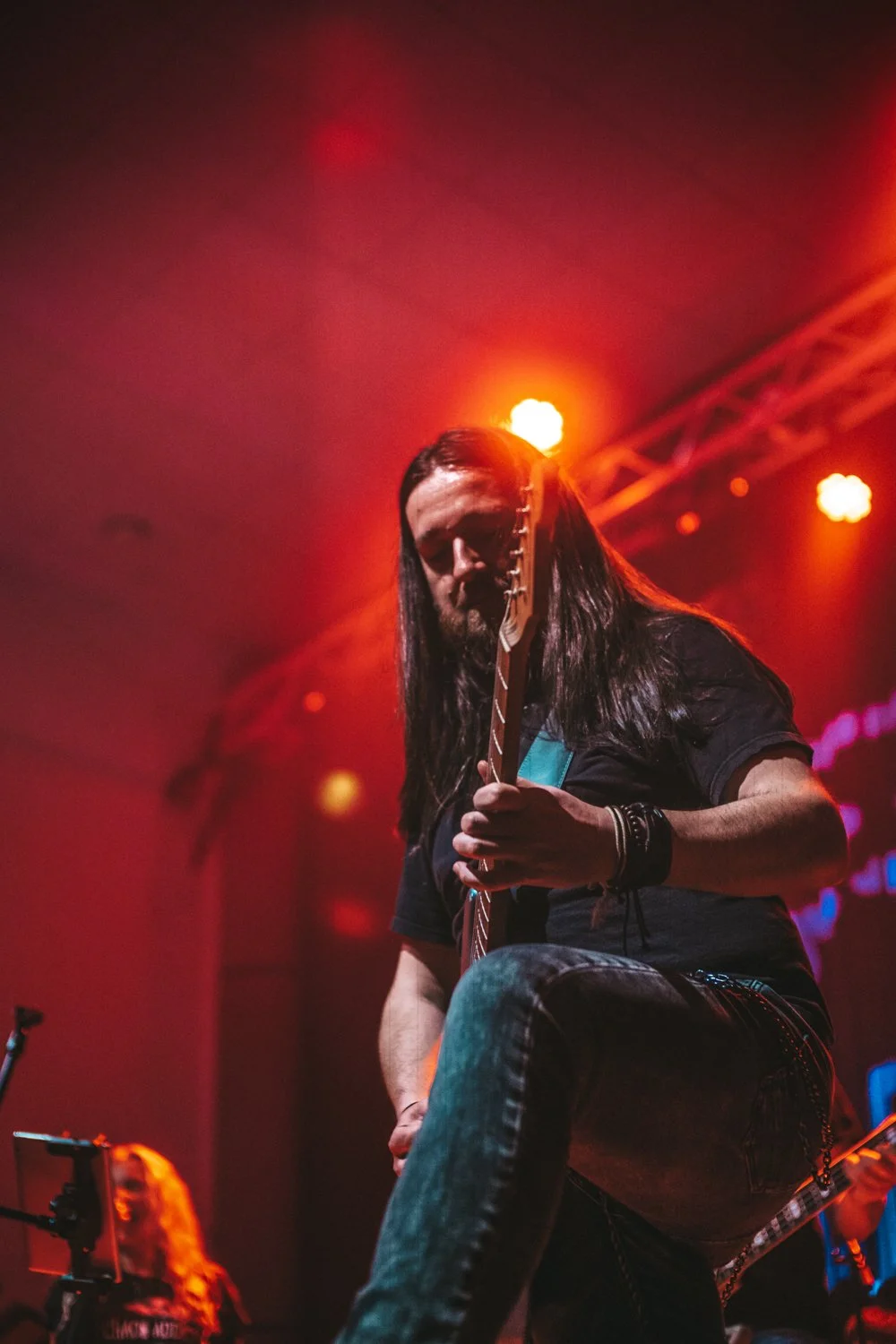 Male guitarist with long hair performing on stage under colorful lights.