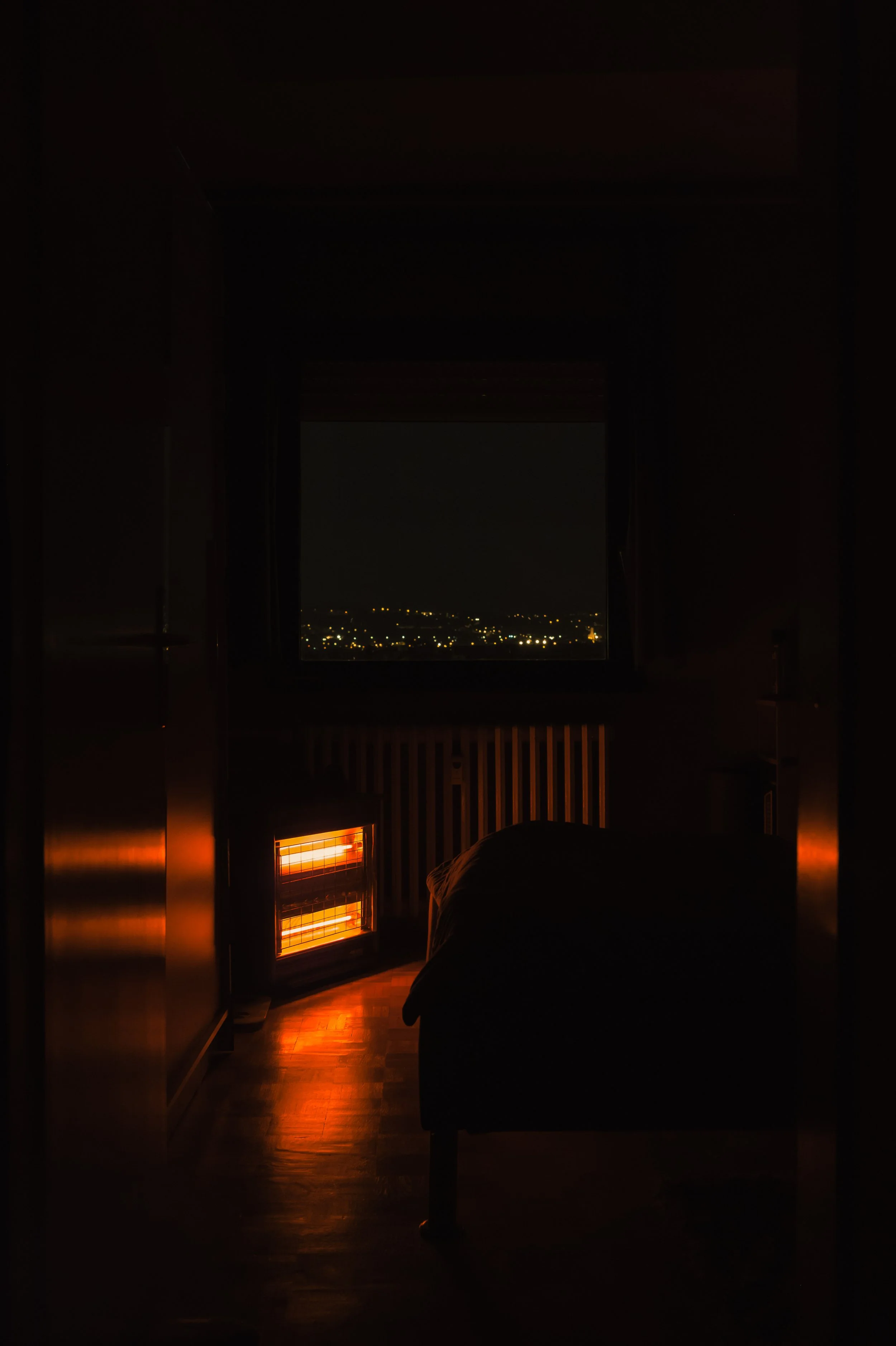Dimly lit bedroom at night with window showing city lights outside, and a space heater glowing orange on the floor.