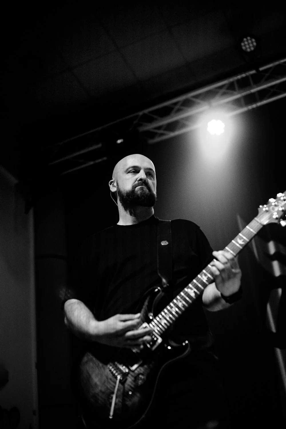 Black-and-white photo of a man with a beard, playing an electric guitar on stage with a bright light shining behind him.