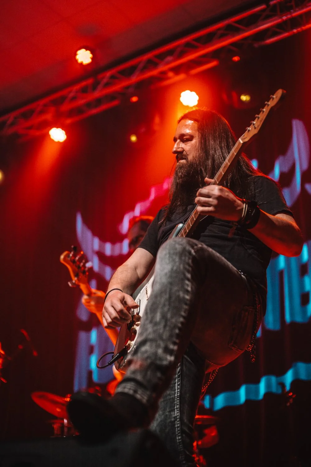 A male guitarist with long hair and a beard performing on stage under red and orange lights, wearing a black t-shirt and jeans.