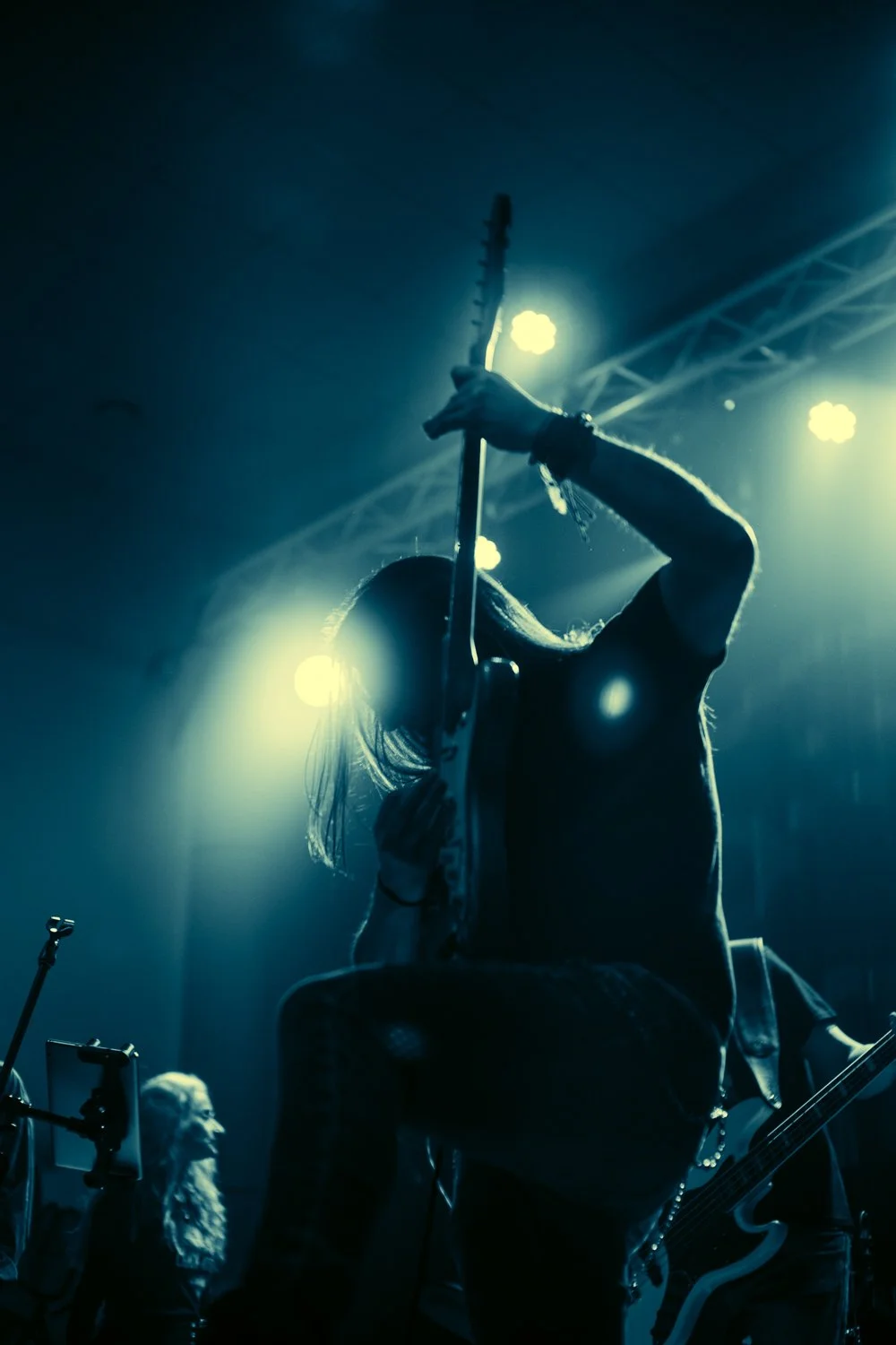 A musician playing an electric guitar on stage, silhouetted against bright stage lights, with another person in the background.