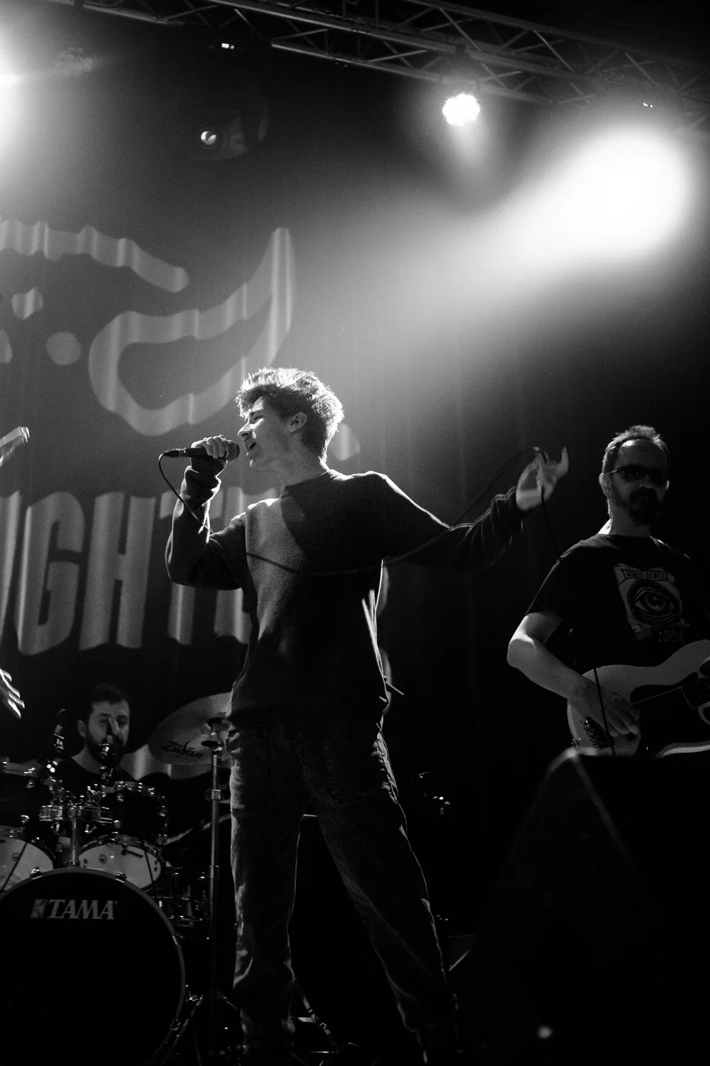 A black-and-white photo of a band performing on stage, with a singer in the center holding a microphone, a drummer in the background, and a guitarist on the right. Bright stage lights illuminate the scene.