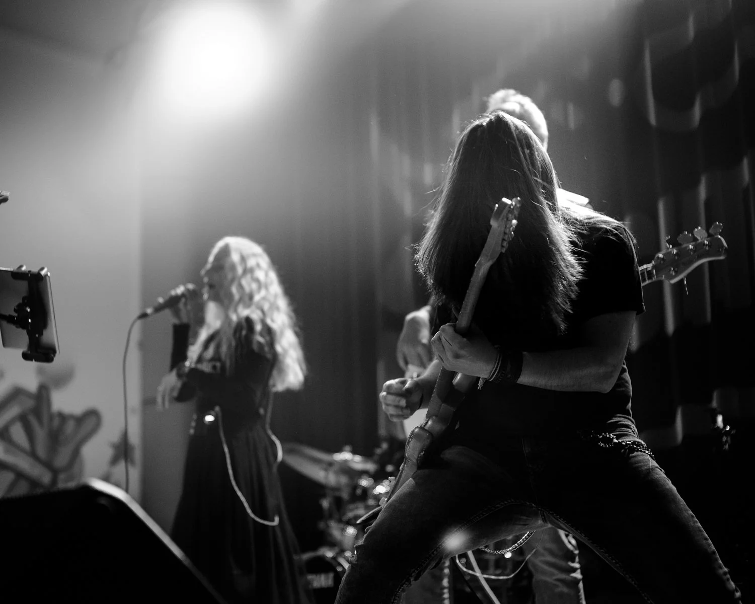 Black and white photo of a band performing on stage, featuring a woman singing into a microphone and a woman playing an electric guitar.
