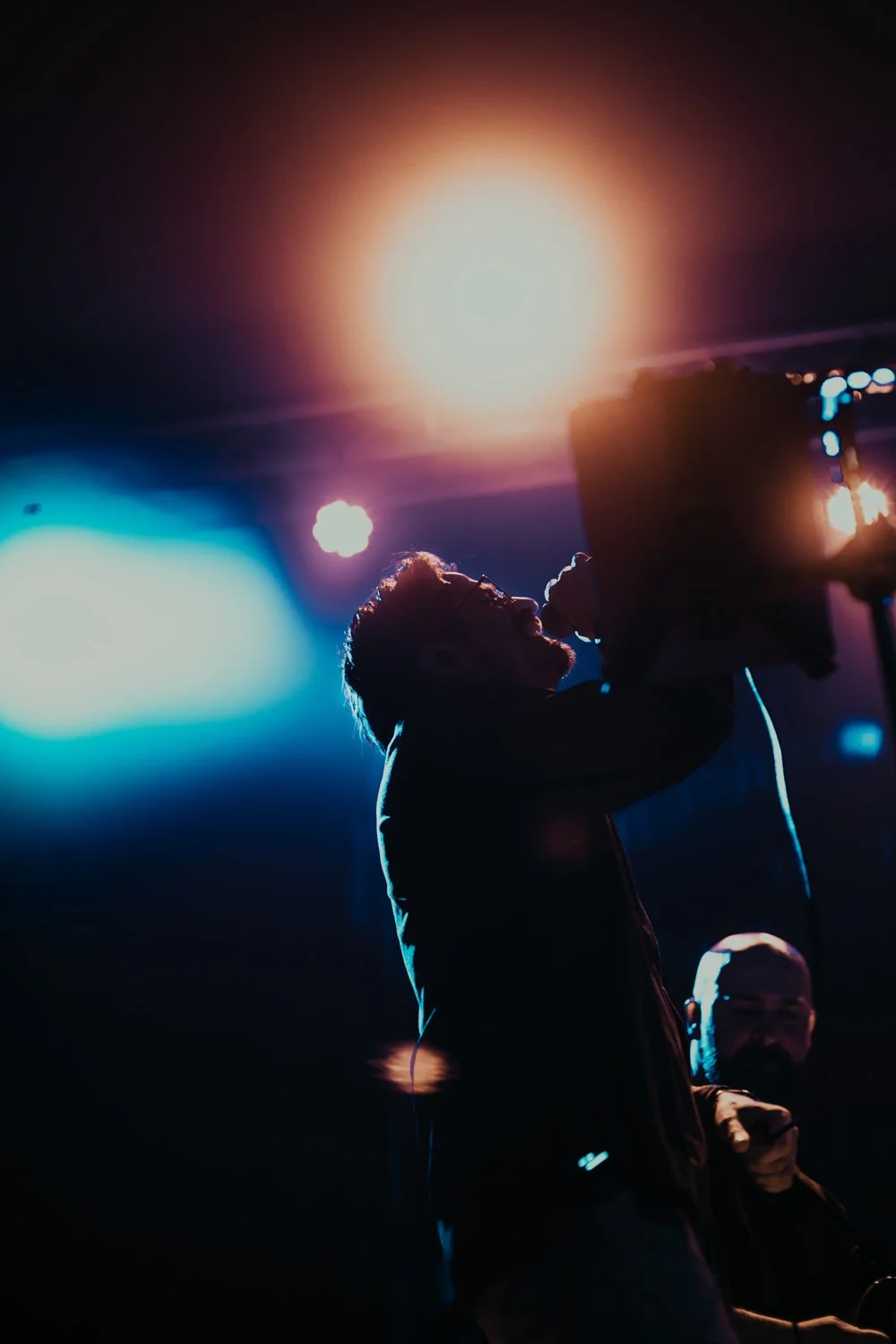 A man with glasses holding a vintage film camera on stage, illuminated by colorful lights, with another person in the background