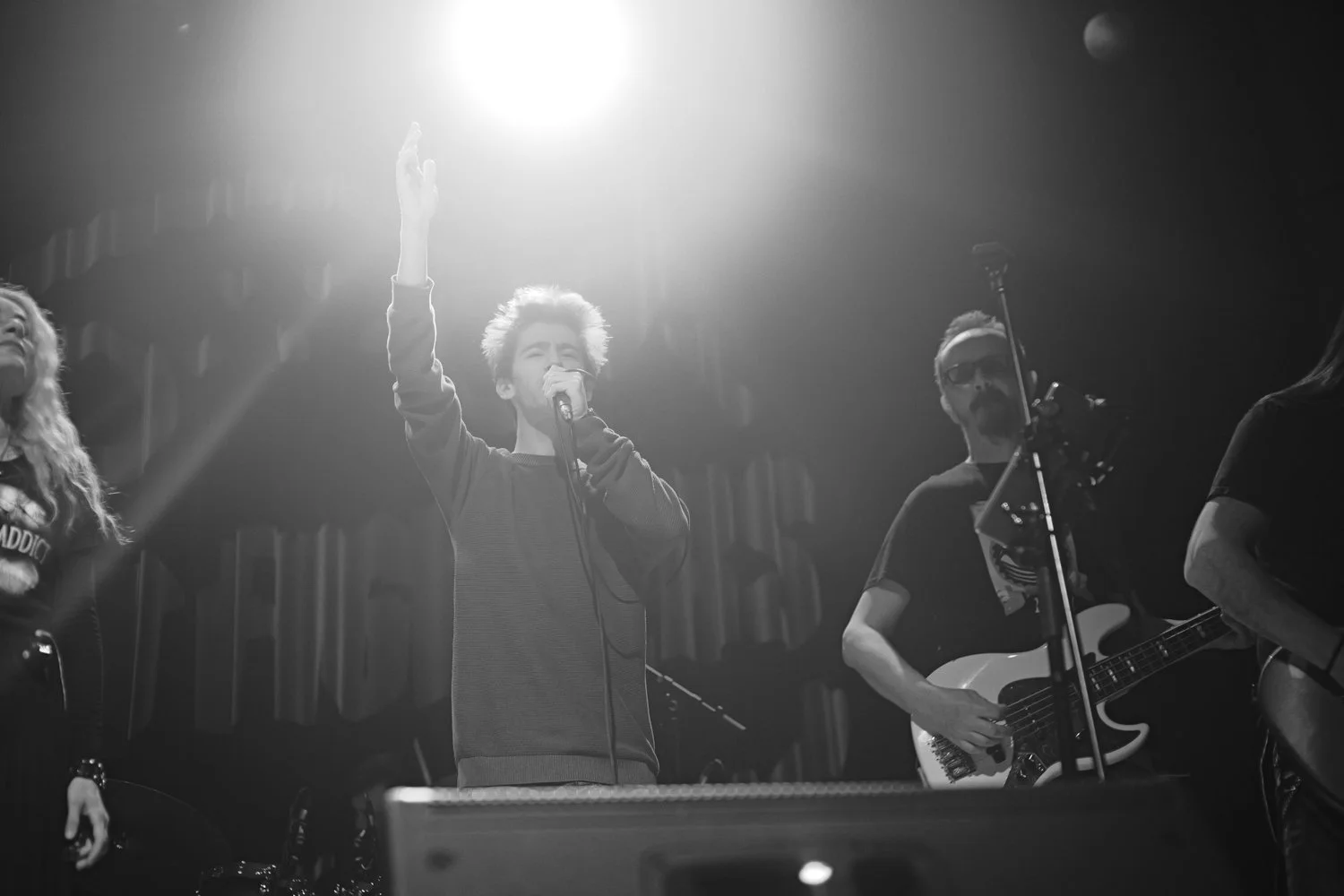 A black and white photo of a musical performance with a singer holding a microphone, raising one hand, and two guitarists standing nearby, one with sunglasses, on a stage with spotlights.