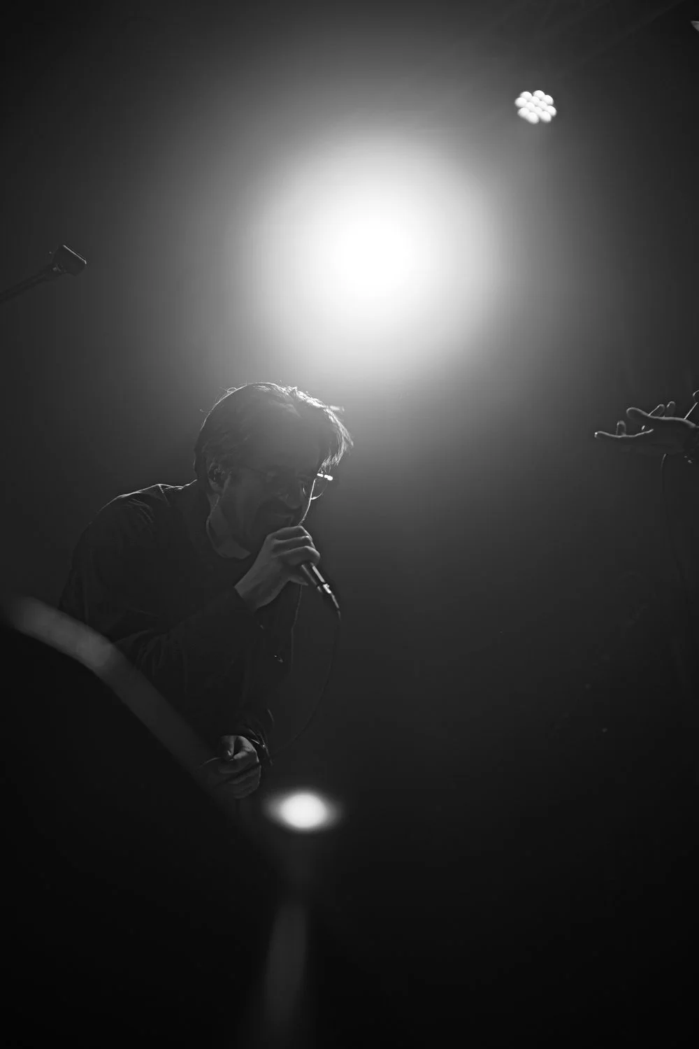 A black and white photo of a man singing into a microphone on stage, illuminated by a bright overhead light with a cloud of small lights in the background.
