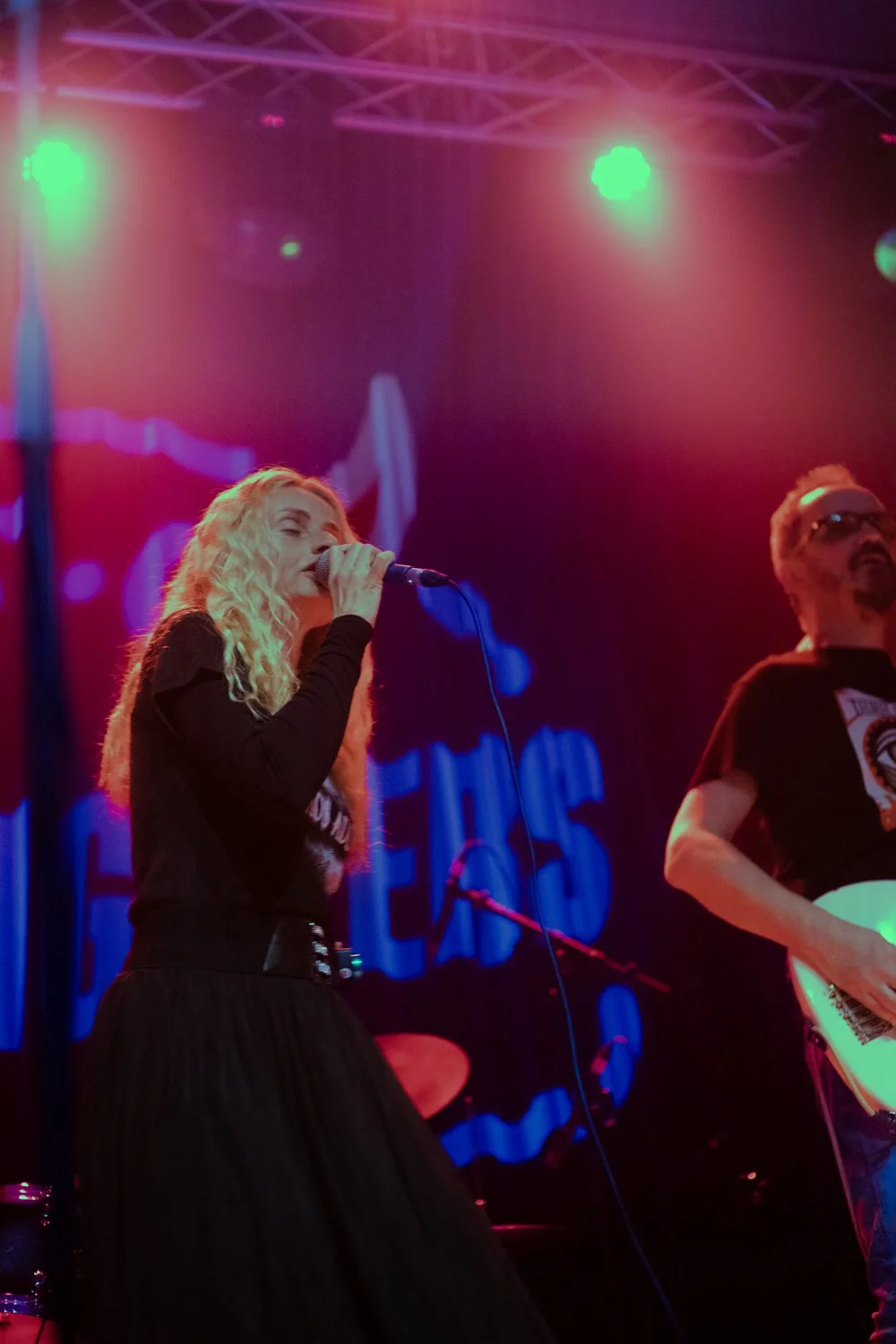 A woman singing into a microphone on stage with colorful lights and a man playing guitar beside her.