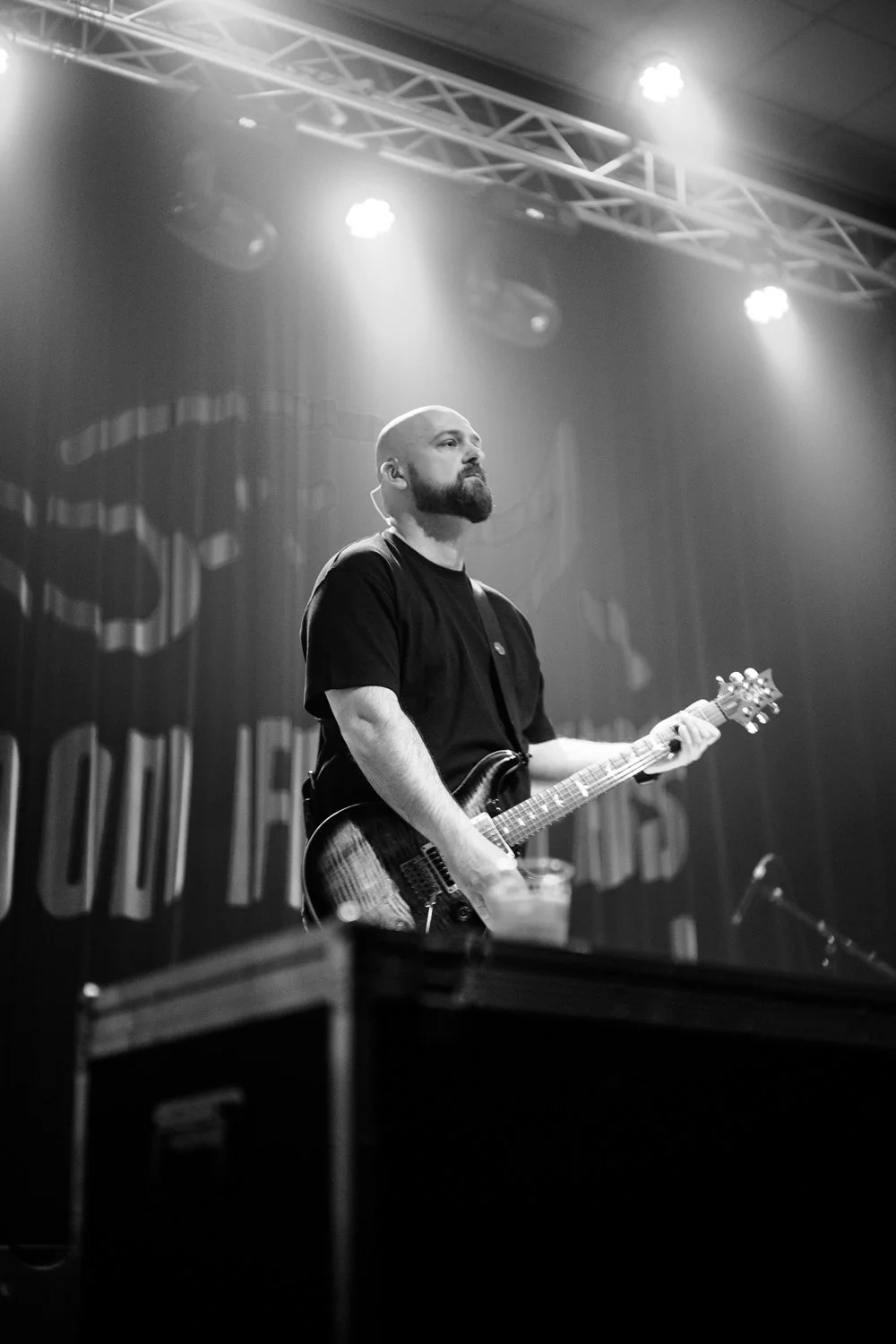 A bearded man playing an electric guitar on stage during a live performance, with stage lighting and a curtain backdrop.