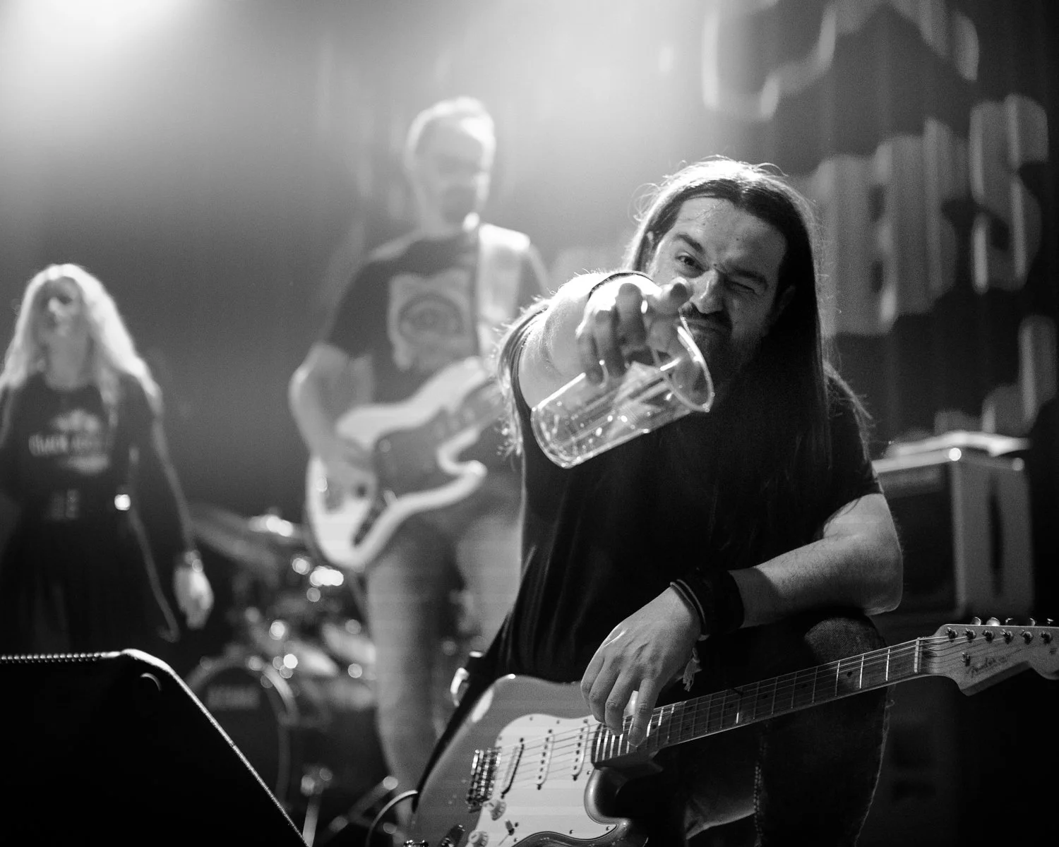 Black and white photo of a man with long hair, holding an electric guitar, smiling and pointing at the camera with a clear plastic cup in his hand, on a stage with band members in the background.
