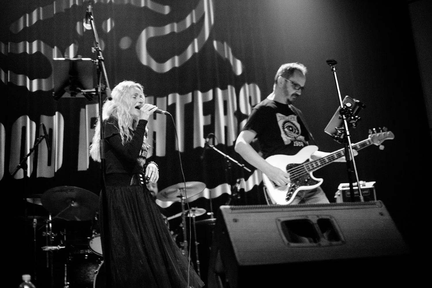 A black and white photo of a band performing on stage, featuring a female singer with long curly hair singing into a microphone, a male guitarist playing an electric guitar, and a drummer in the background. The stage has curtains with a pattern and a
