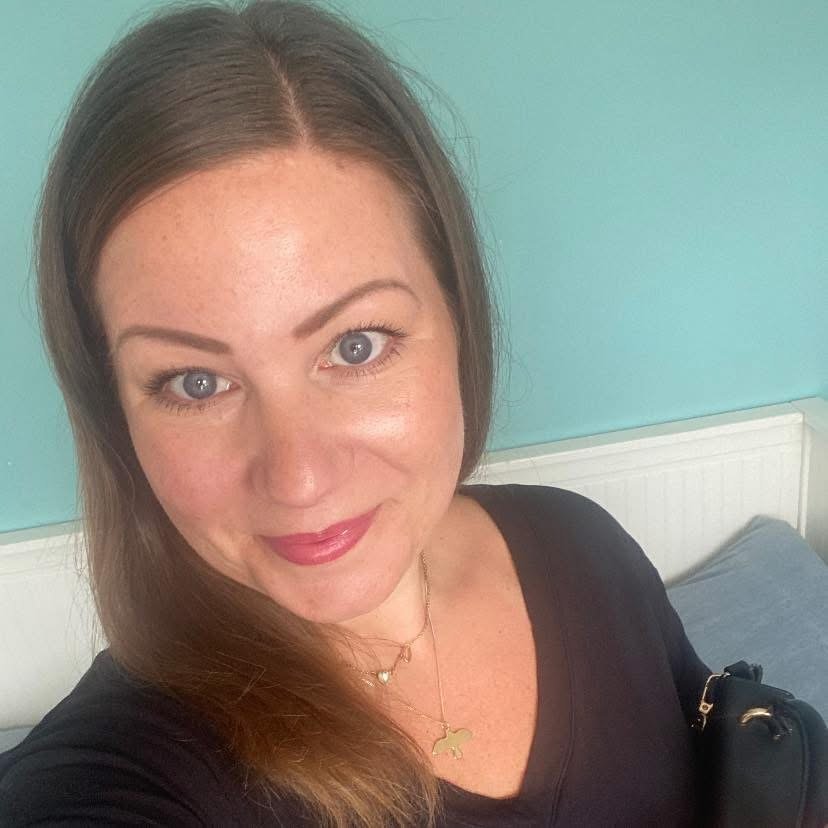 A woman with long brown hair and blue eyes taking a selfie indoors against a teal wall. She is smiling and wearing a black top, layered necklaces, and makeup.