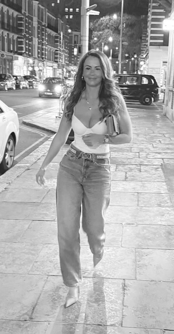 A woman walking on a city sidewalk at night, carrying a clutch, wearing a sleeveless top and jeans, with buildings and cars illuminated in the background.