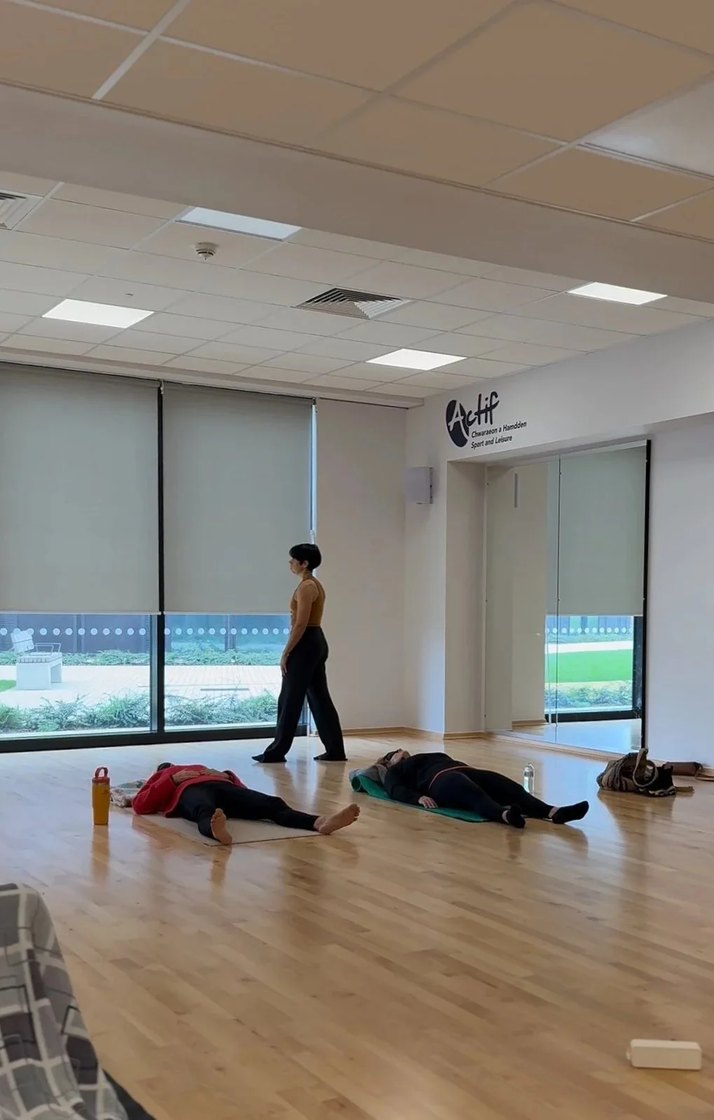 A woman leading a yoga or fitness class in a studio with three participants lying on mats, resting or stretching.