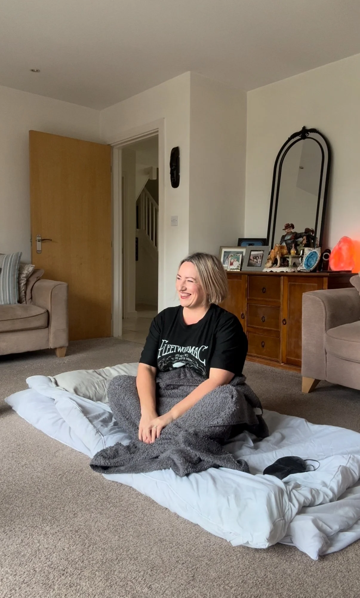 A woman sitting on a mattress on a living room carpet, smiling and wearing a black Fleetwood Mac t-shirt, with a gray blanket on her lap.