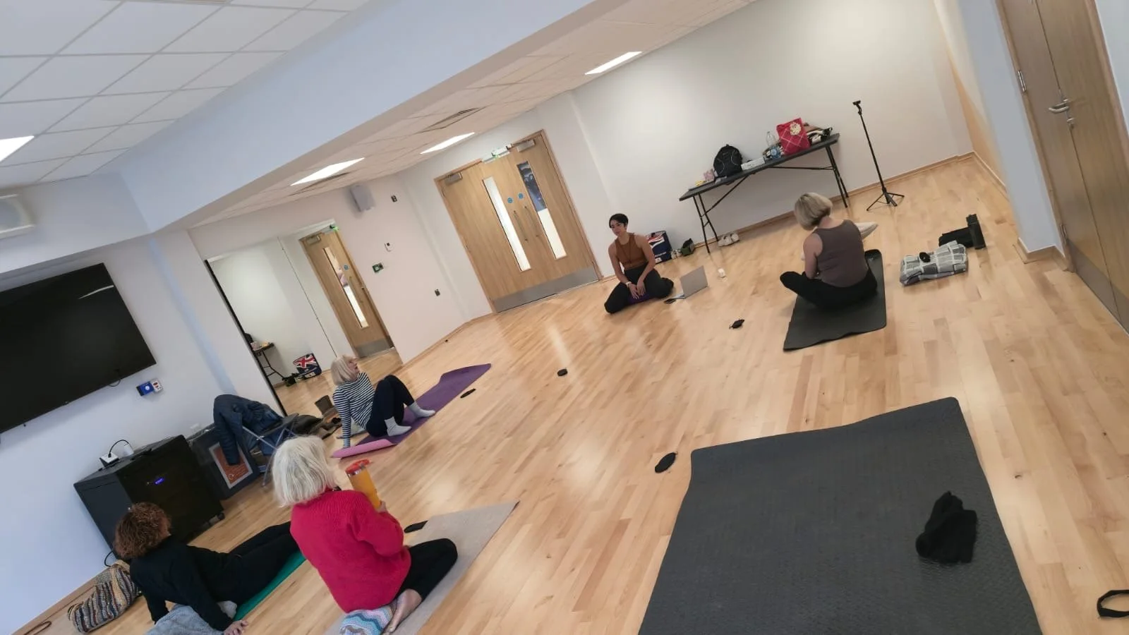 People participating in a yoga class in a spacious, well-lit room with wooden floors, some sitting on yoga mats, engaged in meditation or stretching exercises.