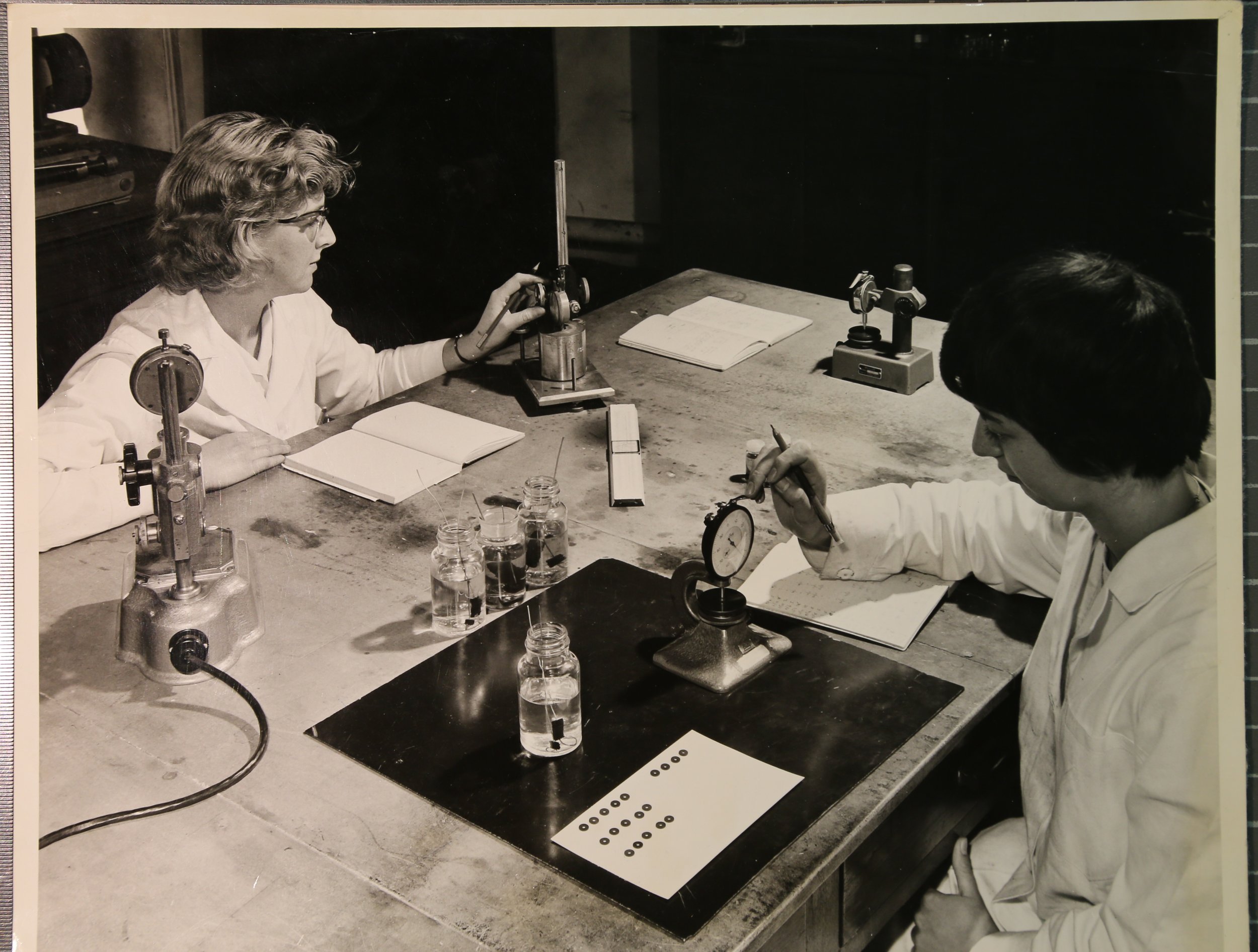 Black-and-white photo of two women working in a laboratory. One woman, with wavy hair and glasses, is using a scientific instrument. The other woman, with darker hair, is examining a small object. The table has various laboratory tools, notebooks, and jars filled with liquids.