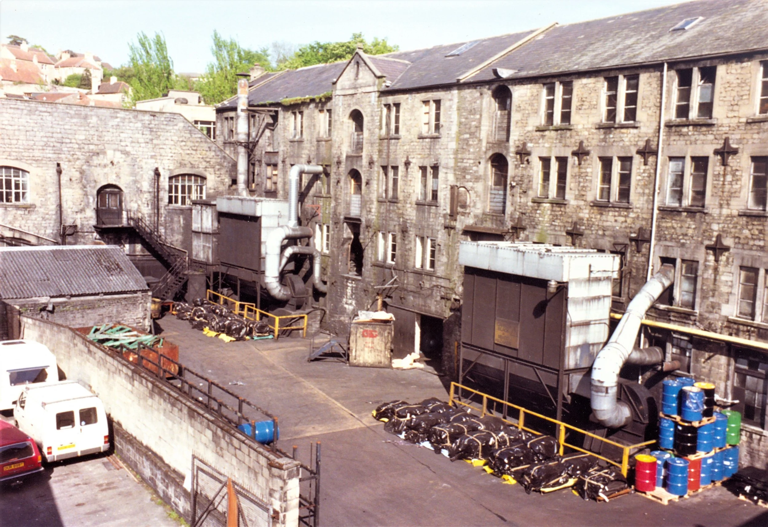 Kingston Mills, Bradford on Avon with multiple windows and a slate roof, industrial pipes and equipment in the foreground, and a parking lot with cars.