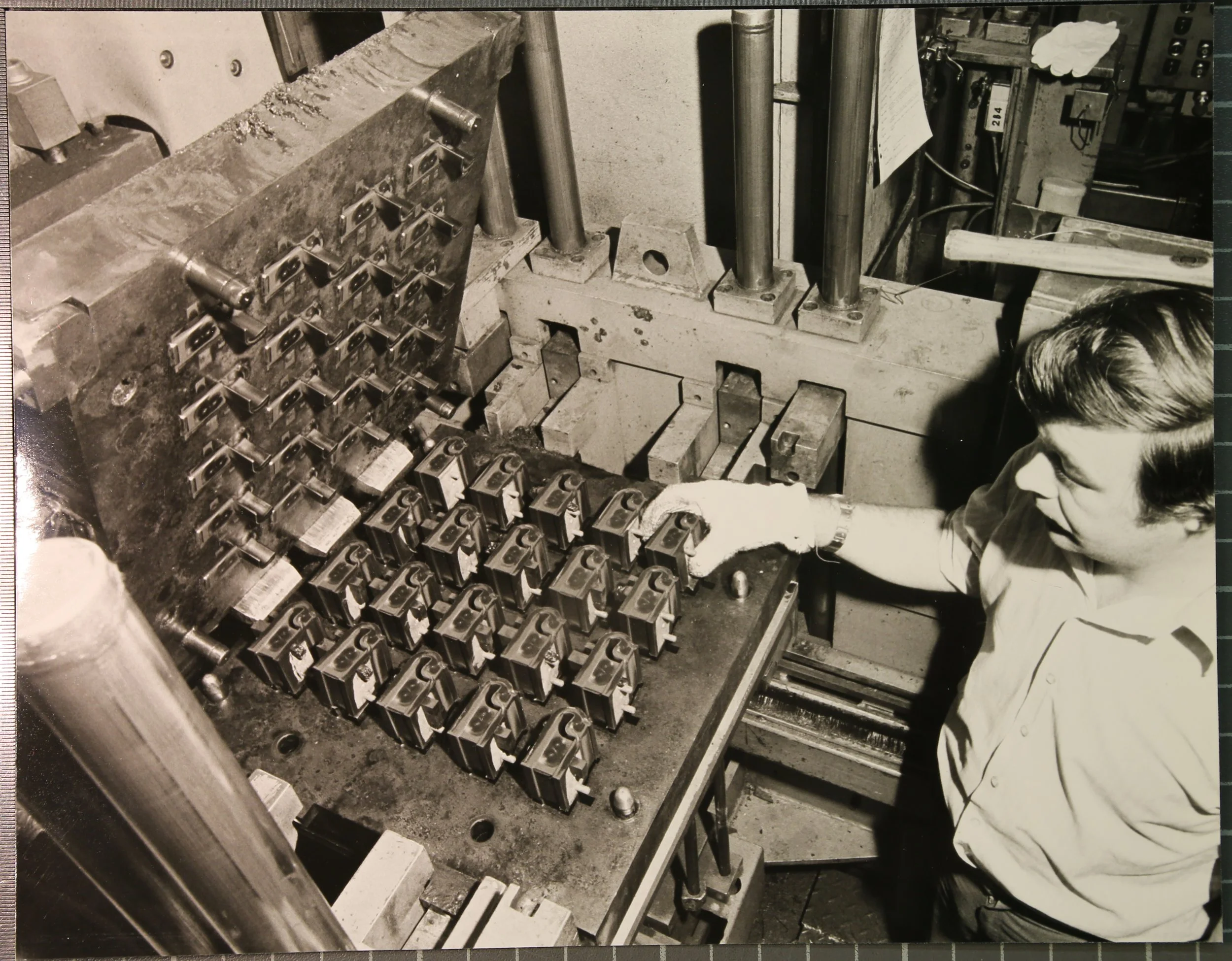 An industrial setting with a worker inspecting a metal component on a machine. The machine has multiple small metal parts arranged in rows, and the worker is looking closely at the assembly.