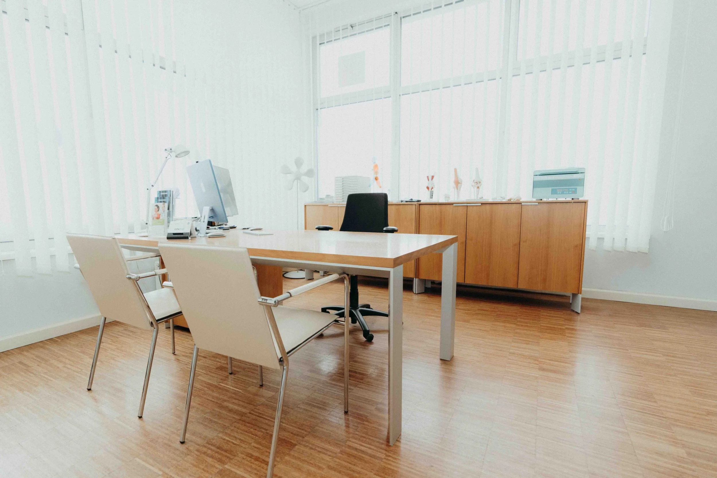 Ein Büro mit einem Schreibtisch, Stühlen, einem Computer, einem Sekretärgerät und einem Holzschrank vor großen Fenstern mit weißen vertikalen Jalousien.
