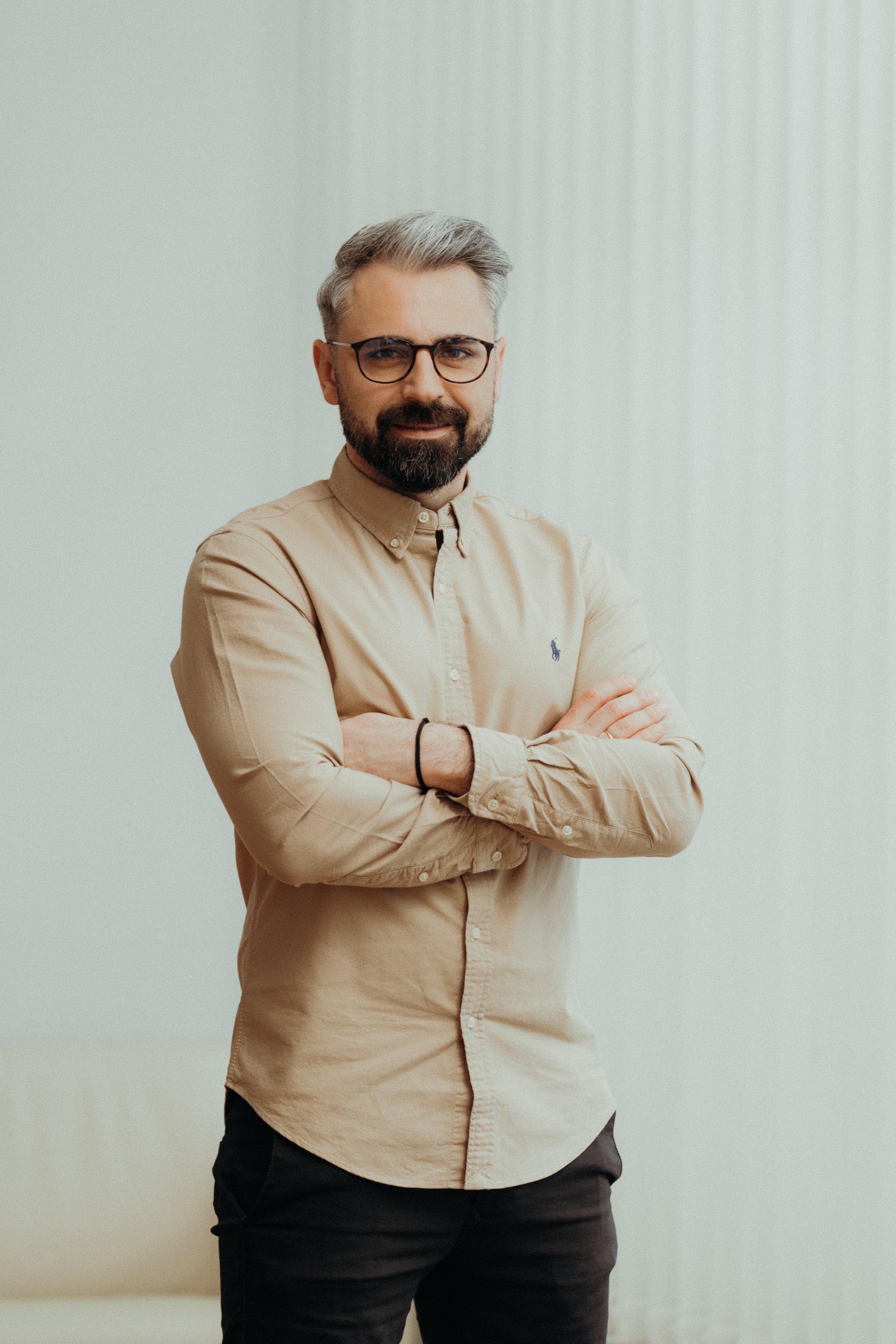 Ein portraitierter Mann mit Brille, Bart und grauem Haar, der lächelnd mit verschränkten Armen steht, vor einem neutralen Hintergrund.