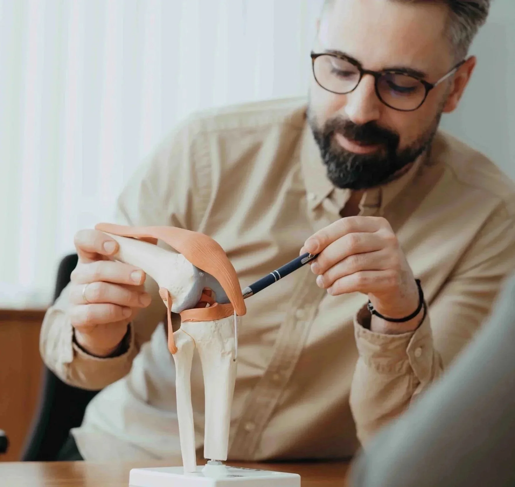 Mann erklärt ein Modell des menschlichen Knies, umgeben von einem Büro, trägt Brille und beige Hemd.