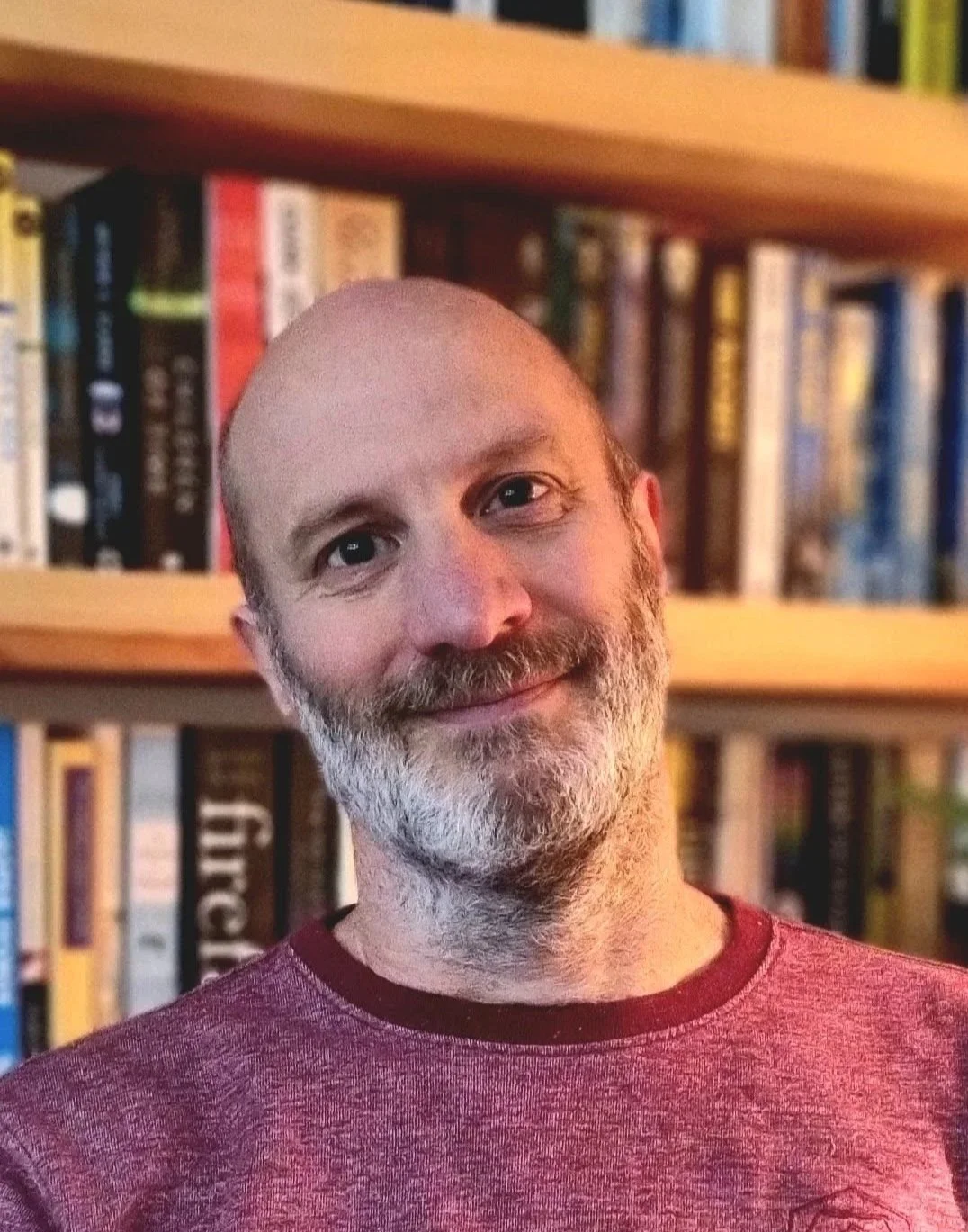 A man with a beard and a friendly smile stands in front of a bookshelf filled with books.